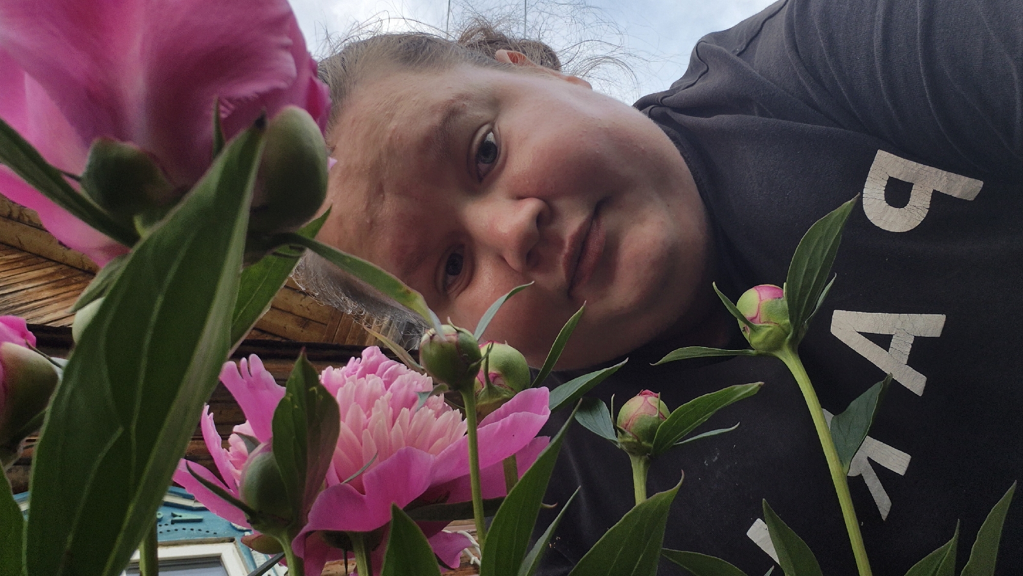 This is a low-angle, close-up, selfie-style photograph featuring a person and pink peonies. The person, who appears to be an adult, is positioned in the upper right quadrant of the frame, with their face tilted towards the camera, looking directly at the viewer. Their hair is pulled back, and they are wearing a dark t-shirt with large, light-colored, cracked letters that read "BA". The face of the person is illuminated by natural light, suggesting it is daytime. The background consists of a wooden structure, likely a building, and hints of a light-colored sky, indicating it could be overcast or late afternoon.
In the foreground, several vibrant pink peonies are prominently displayed. One large, fully bloomed peony is on the left, partially obscuring the person's face. Smaller buds and partially opened blooms are scattered throughout the frame, adding depth and color to the composition. The lush green leaves and stems of the peony plants are also visible, creating a natural, garden-like setting. The overall mood of the image is intimate and personal, with the close proximity of the flowers and the direct gaze of the subject. The setting appears to be outdoors, possibly in a rural area given the wooden structure and abundant flowers, consistent with the provided location context of Дойная, Russia.