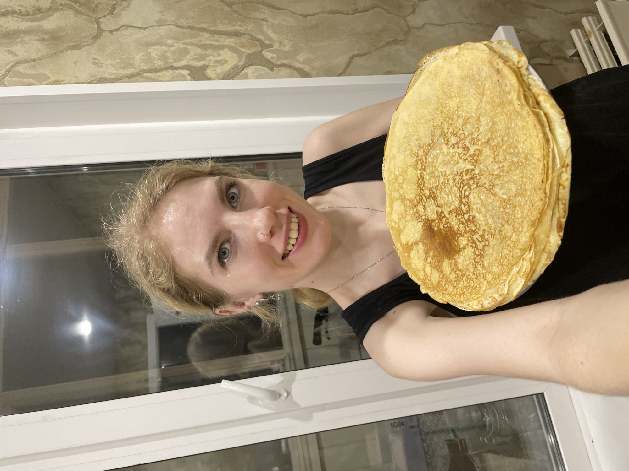 The scene is located indoors, within a residential dwelling in Izhevsk, Russia. A young adult female, with blonde hair pulled back, light complexion, and blue eyes, is positioned centrally and smiling directly at the viewer. She is wearing a black tank top and a silver-colored chain necklace. Her left ear exhibits multiple piercings including hoops and studs, one with a small turquoise bead, and her right ear has a single stud. A small stud is visible on her left nostril. She is holding a large stack of approximately 15-20 golden-brown, circular pancakes (blini) on a white plate, positioned in front of her lower chest. Her left forearm is partially visible, supporting the plate. The background features a white PVC window frame on the left, with a white handle. Through the window, a dark exterior suggests nighttime, and a distinct reflection of an interior light source is visible. To the right of the window, a wall is covered with light beige wallpaper featuring an abstract, textured pattern. Below the window, a white windowsill and a section of a white radiator are visible.