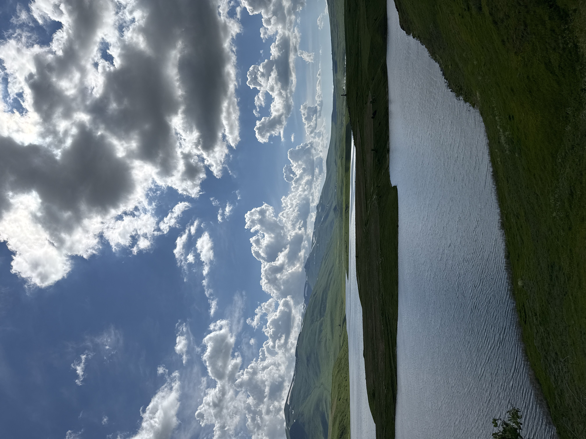 The image depicts a serene landscape with a large body of water in the foreground, reflecting the dramatic, cloud-filled sky. Rolling green hills, some with patches of snow, stretch across the horizon. The sky is a vibrant blue, punctuated by numerous fluffy white clouds and darker, more dramatic storm clouds, creating a play of light and shadow across the scene. A dark green, grassy slope dominates the right foreground, meeting the edge of the water. The scene is devoid of any people or structures, suggesting a remote, natural environment. Based on the description, this landscape could be the vicinity of Tsghuk, Armenia, known for its natural beauty and mountainous terrain.
