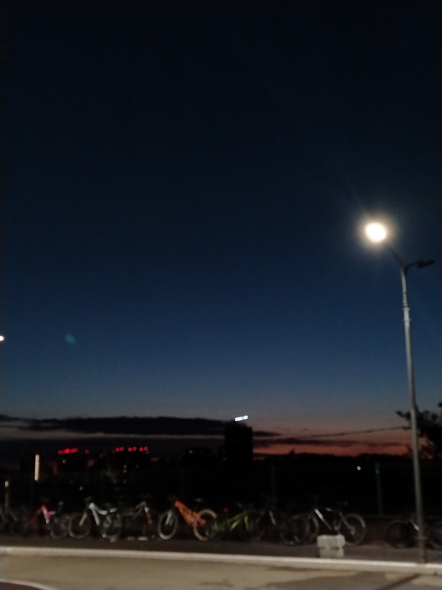 The image shows a row of bicycles parked along a sidewalk in Izhevsk, Russia at dusk. A streetlight illuminates the right side of the frame. In the background, the city skyline is visible with several lit buildings and streetlights. The sky is dark blue with a hint of orange near the horizon.