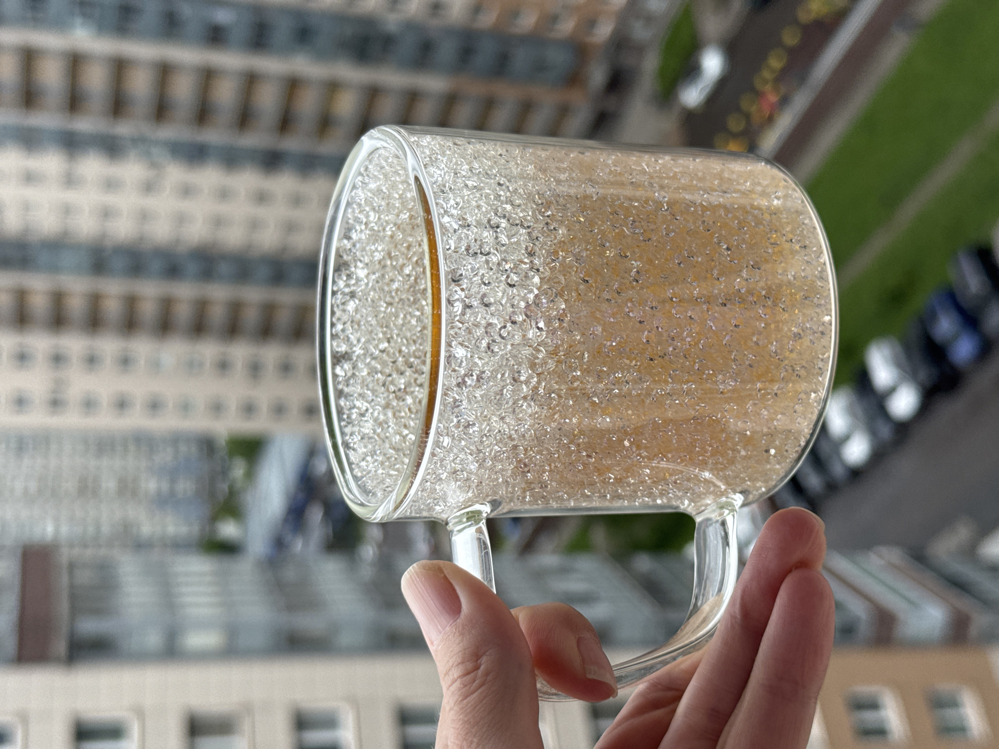 The image shows a transparent glass mug, partially filled with a yellowish liquid, held by a human hand. The liquid appears to have small, translucent beads suspended within it. The background is a blurred cityscape, featuring buildings and a street with parked and moving vehicles. The location is in a city, but the specific city is unknown. The perspective suggests the photo was taken from a high vantage point, looking down at the city.