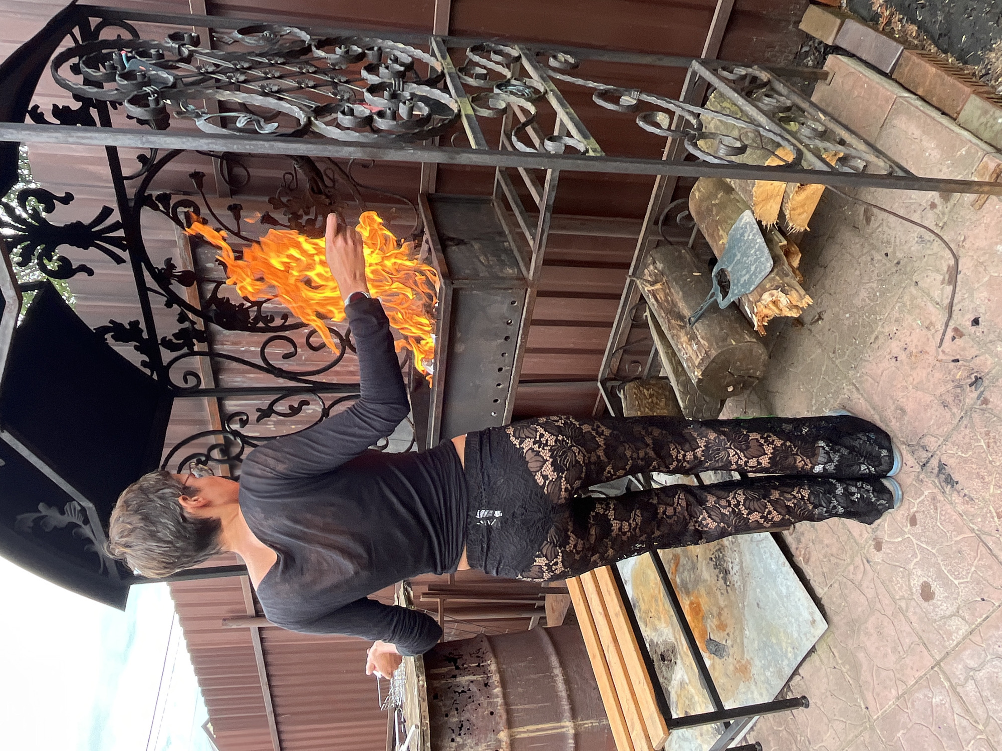 A person with short, dark hair and wearing a black long-sleeved shirt and black lace pants stands in front of an outdoor grill with flames. The person is holding something towards the flames. To the left of the grill is a wooden bench, and to the right are logs and a metal grate. The background consists of a brown fence and a cloudy sky. The scene appears to be in a backyard or outdoor cooking area in the village of Veljaminovo, Russia.