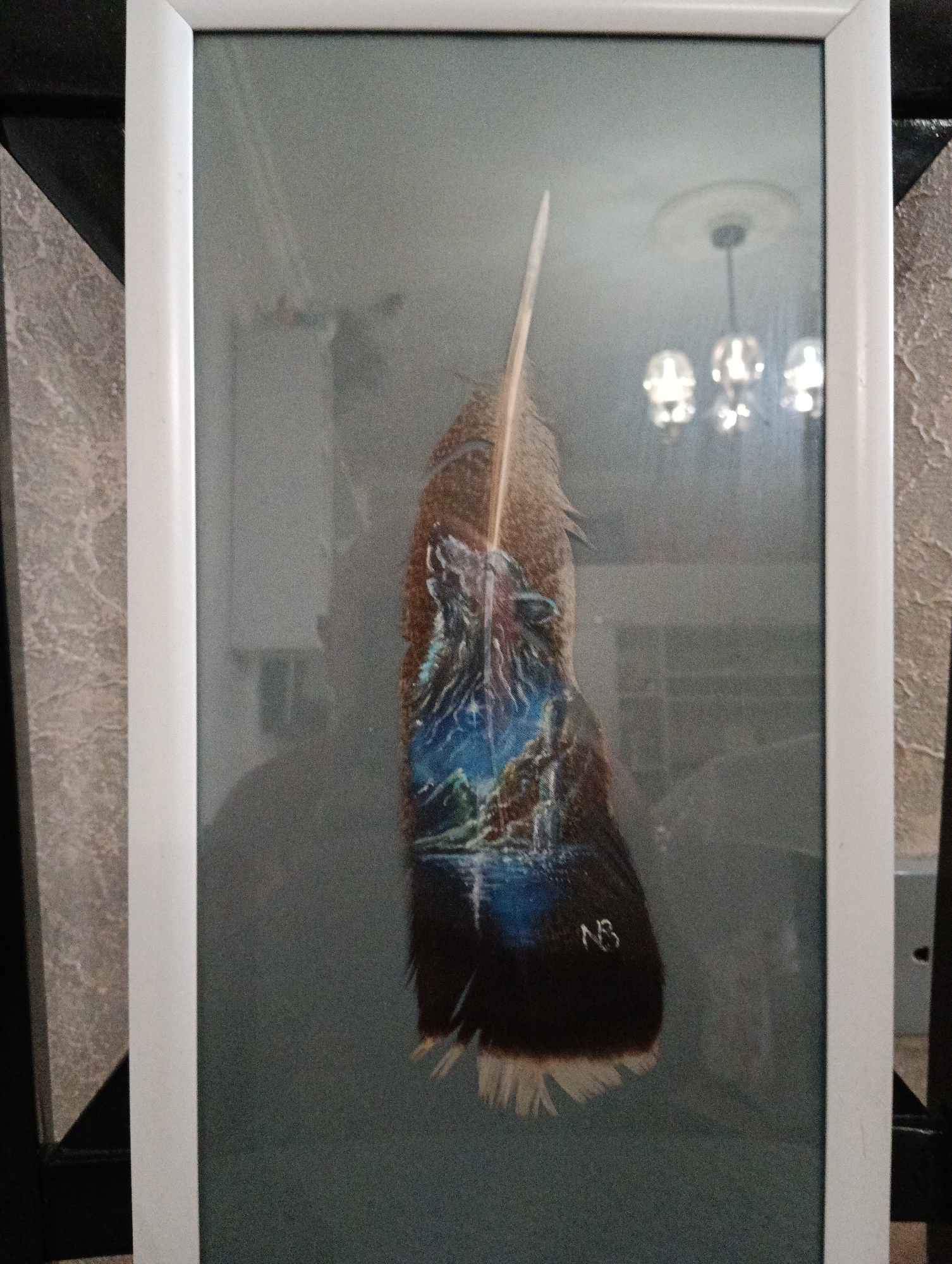 A framed artwork featuring a painted feather is displayed against a textured wall. The feather, oriented vertically, is adorned with a painted scene depicting a wolf howling at the moon amidst a mountainous landscape with a blue sky. The artist's initials, "NB," are visible on the lower portion of the feather. In the background, a reflection shows a chandelier hanging from a ceiling and other interior elements, suggesting the artwork is displayed indoors. The location is a residential or gallery setting in the city of Baku, Azerbaijan.