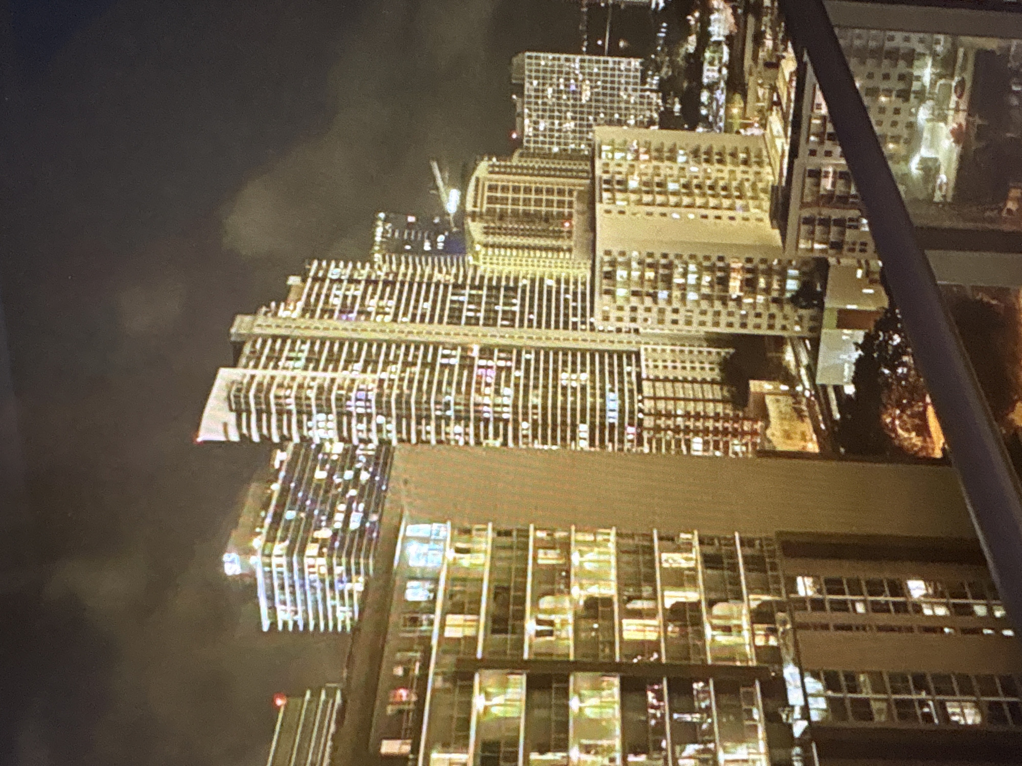 The image depicts a nighttime cityscape characterized by numerous illuminated high-rise buildings. The buildings are densely packed, suggesting an urban environment. Many of the structures have a modern architectural style, with a significant number of windows visible, each glowing with interior light. The sky above is dark and partially obscured by clouds, which are subtly illuminated by the city lights below. A balcony railing runs across the foreground of the image, indicating the perspective is from an elevated vantage point, likely a residential unit or a higher floor of another building. The scene is devoid of visible people. The overall impression is one of a bustling, well-lit city at night. The question specifies the location as Pori, Finland, but the visual characteristics do not specifically identify it as such.