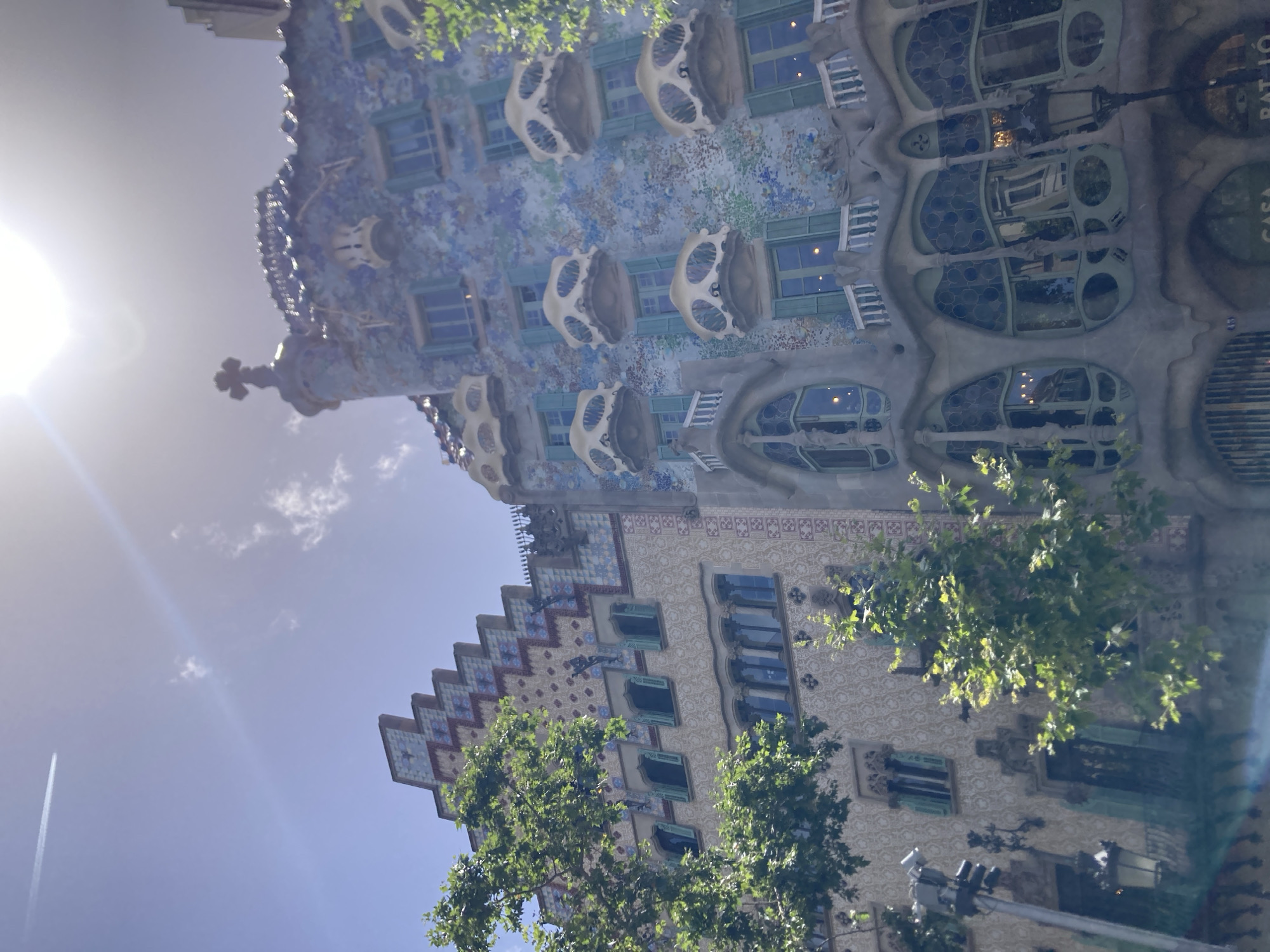 This is a downward-angled photograph of Casa Batlló in Barcelona, Spain. The building's unique facade, characterized by its organic, bone-like balconies and mosaic-tiled surfaces, is prominently featured. Green trees with dense foliage partially obscure the lower sections of the building, suggesting a daytime setting. The sky above is a bright blue, with a few scattered white clouds and a prominent lens flare from the sun, indicating it is a sunny day.
The architecture itself is the primary subject, showcasing intricate details like the patterned tiles, the rounded window frames, and the decorative elements that give the building a whimsical and surreal appearance. The facade is a riot of color, with blues, greens, and other vibrant hues, suggesting the lively character of the city.
While there are no people visible in the frame, the building itself, with its distinct architectural style, serves as a powerful representation of Antoni Gaudí's work and the vibrant cultural landscape of Barcelona. The overall impression is one of artistic brilliance and a harmonious blend of nature and architecture. Visible text includes "CASA" and "BATLLÓ" on the building itself.