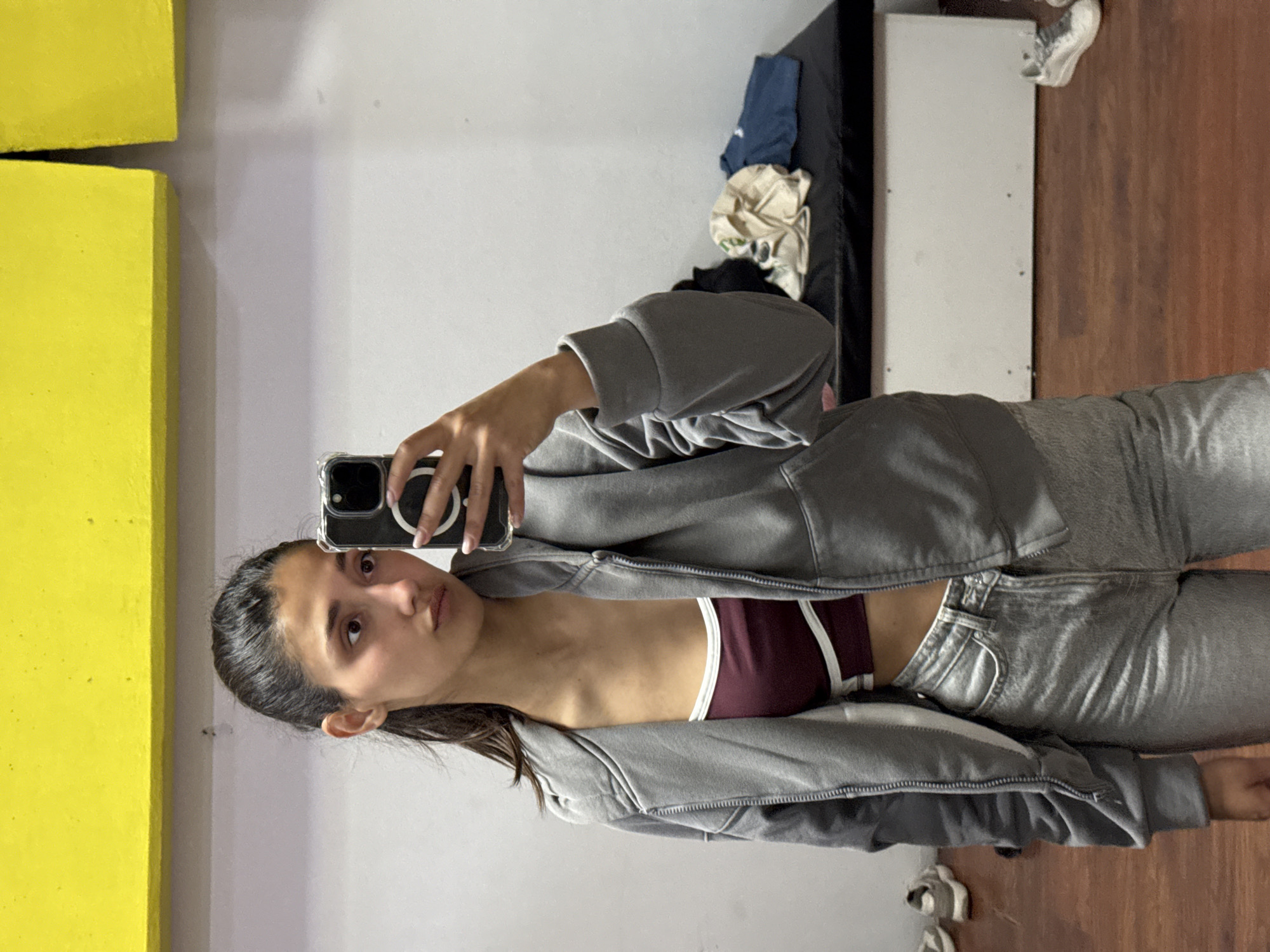 A young woman is taking a selfie in a room with a combination of gray and yellow walls. She is wearing a gray zip-up hoodie over a burgundy sports bra with white accents, and light gray jeans. The room contains a black padded surface along a wall, a white shelf-like structure, and a wooden floor. The location is unidentifiable as Bar, Montenegro based on this image alone.