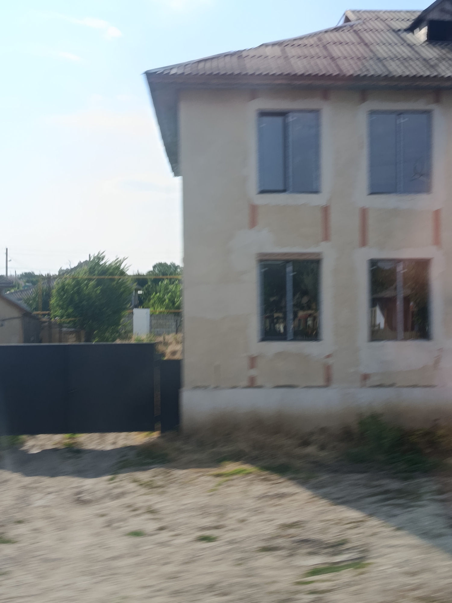 The image depicts a two-story house with a light-colored facade, situated in Chirsova, Moldova. The house has a grey, corrugated roof and four large windows on the visible facade, two on each floor. The windows on the upper floor are reflective, showing a clear sky and some clouds. The windows on the lower floor are also reflective, showing the interior of the house, which appears to be furnished with tables and chairs. The house is partially under construction or renovation, as indicated by the exposed red brick-like elements around the window frames.
In the foreground, a dark, solid metal gate runs across the lower left of the image. Beyond the gate, a sandy area with sparse green vegetation is visible. In the background, there are trees, other houses with corrugated roofs, and a telephone pole. The overall environment suggests a semi-rural or suburban setting. The lighting indicates it is daytime, with bright sunlight casting shadows. The image is slightly blurred, possibly due to motion during capture.