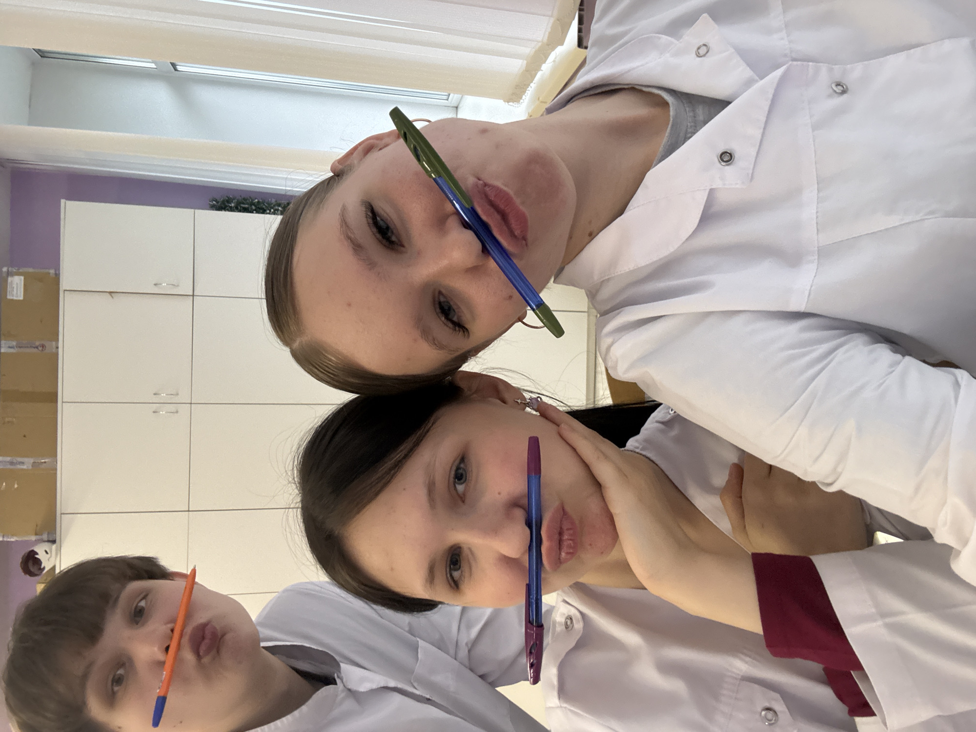 Here is a description of the image:
Three individuals are posing for a photo. All three individuals are holding pens horizontally between their noses and upper lips. The individuals are wearing what appears to be white lab coats. The background includes white cabinets and a window with blinds. The location is unspecified, but presumed to be indoors. The scene depicts a lighthearted moment of individuals posing with pens. Based on facial features, it is not possible to identify ethnicity, but the subjects appear to be young adults. The photo shows a candid moment.