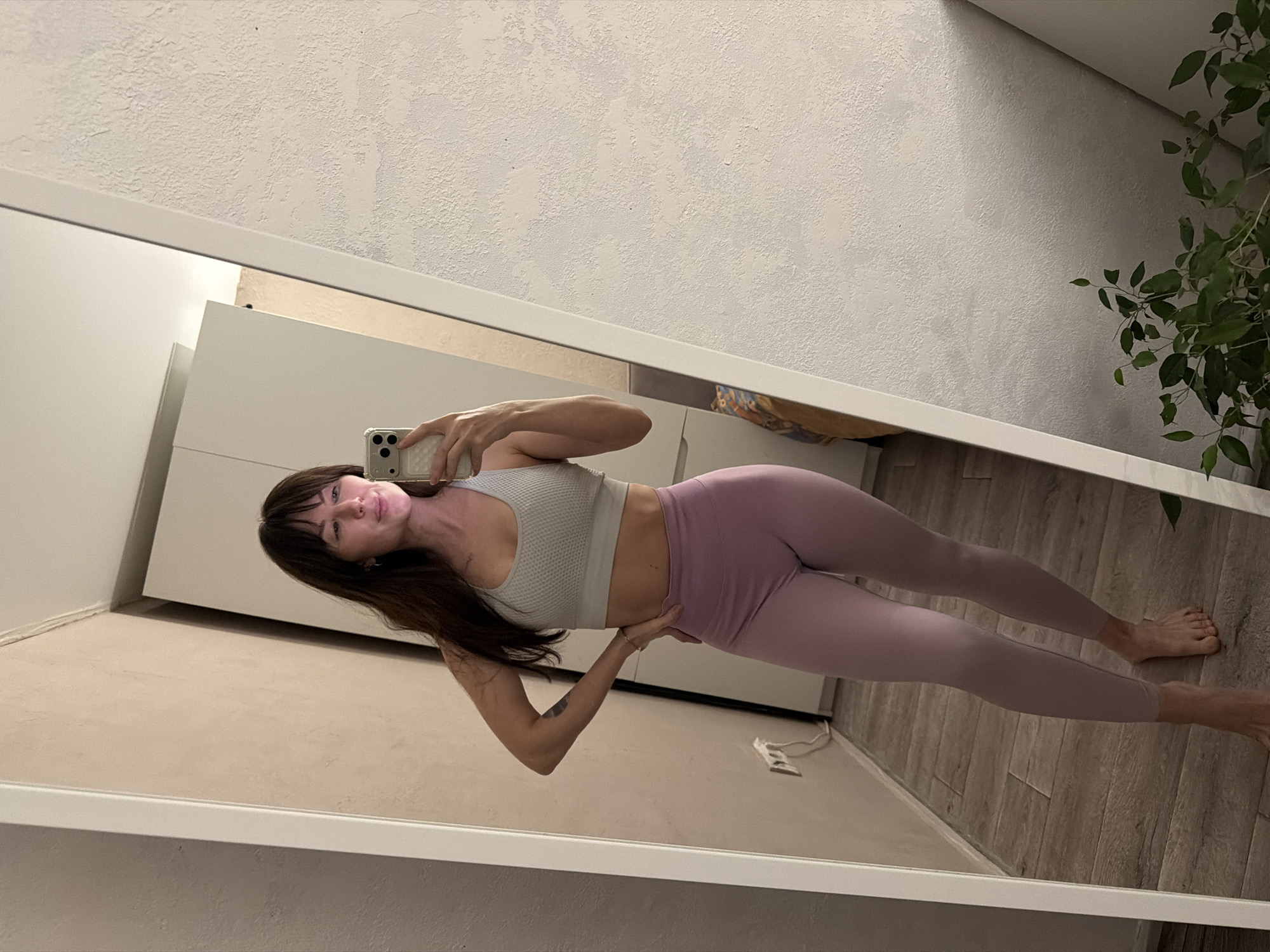 The image displays a mirror selfie of a woman taken indoors, in what is contextually identified as Perm, Russia. The woman has long dark hair with bangs and is wearing a light gray athletic crop top and light purple leggings. She holds a smartphone with a light-colored, textured case in her right hand, capturing her reflection. Her left hand rests on her hip, contributing to her posed stance. Her expression is calm with a slight, subtle smile, and her bare feet are visible towards the bottom of the frame.
The background, seen through the mirror's reflection, consists of light-colored, textured walls. To the left, white or light-toned furniture, possibly a cabinet or wardrobe, is visible. On the right, a portion of a green leafy plant is reflected. The floor appears to be made of wood or laminate, and a white electrical cord is visible on it. The lighting suggests a well-lit indoor environment, possibly daytime. The image itself is rotated to a landscape orientation, though the selfie subject is depicted upright in the reflection. There is no visible text in the image.