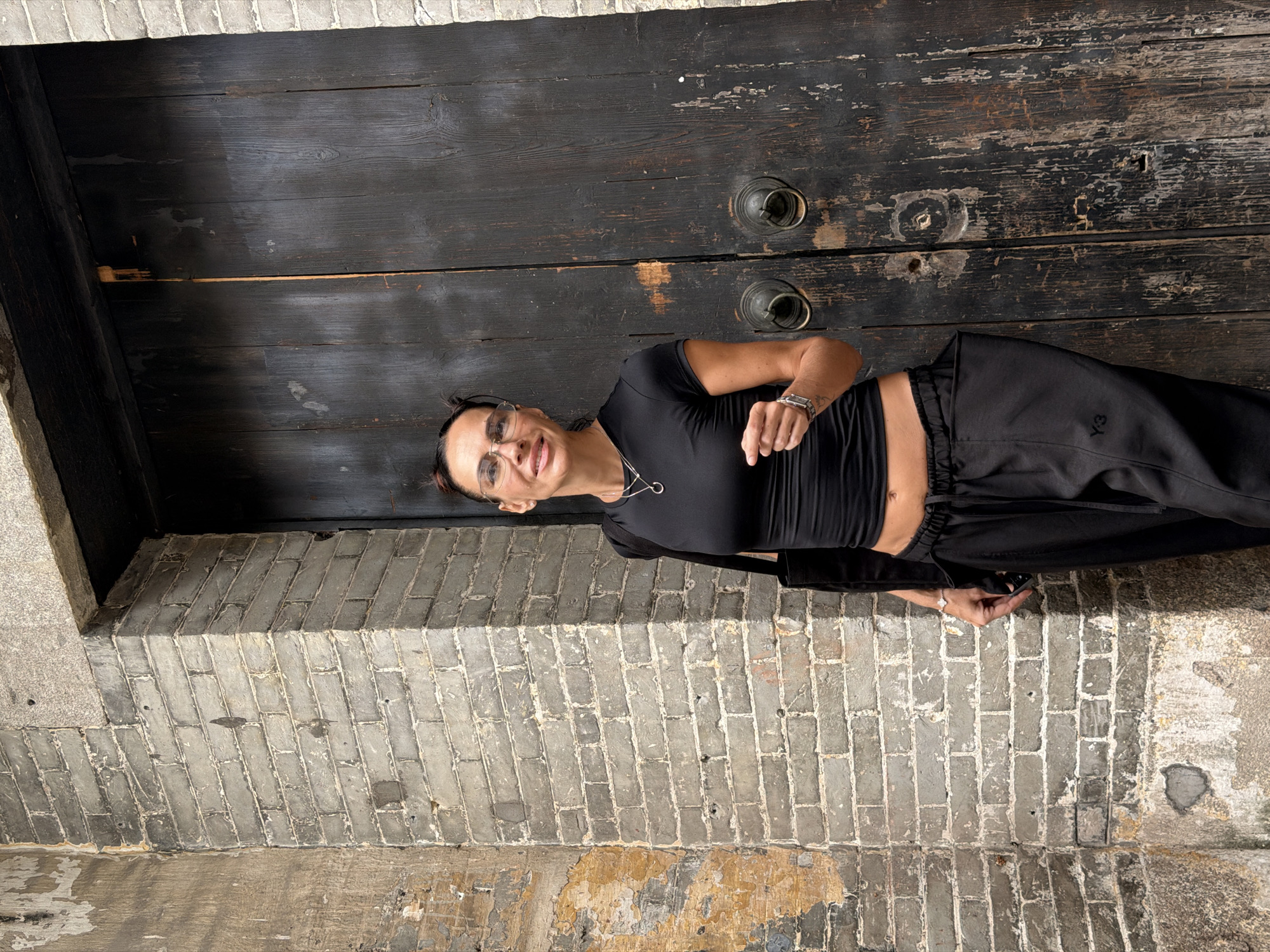 A medium shot shows a woman with dark hair pulled back, wearing clear-rimmed glasses, a black crop top, and black Y-3 sweatpants. She stands in front of a large, dark wooden door with two ornate brass handles. To her left is a rough, grey brick wall with patches of peeling yellow plaster. Her left arm is bent, and her hand is open, gesturing slightly. She carries a black tote bag over her left shoulder. The setting appears to be an outdoor alleyway in the Chancheng District of China, characterized by its aged brickwork and weathered wooden door.