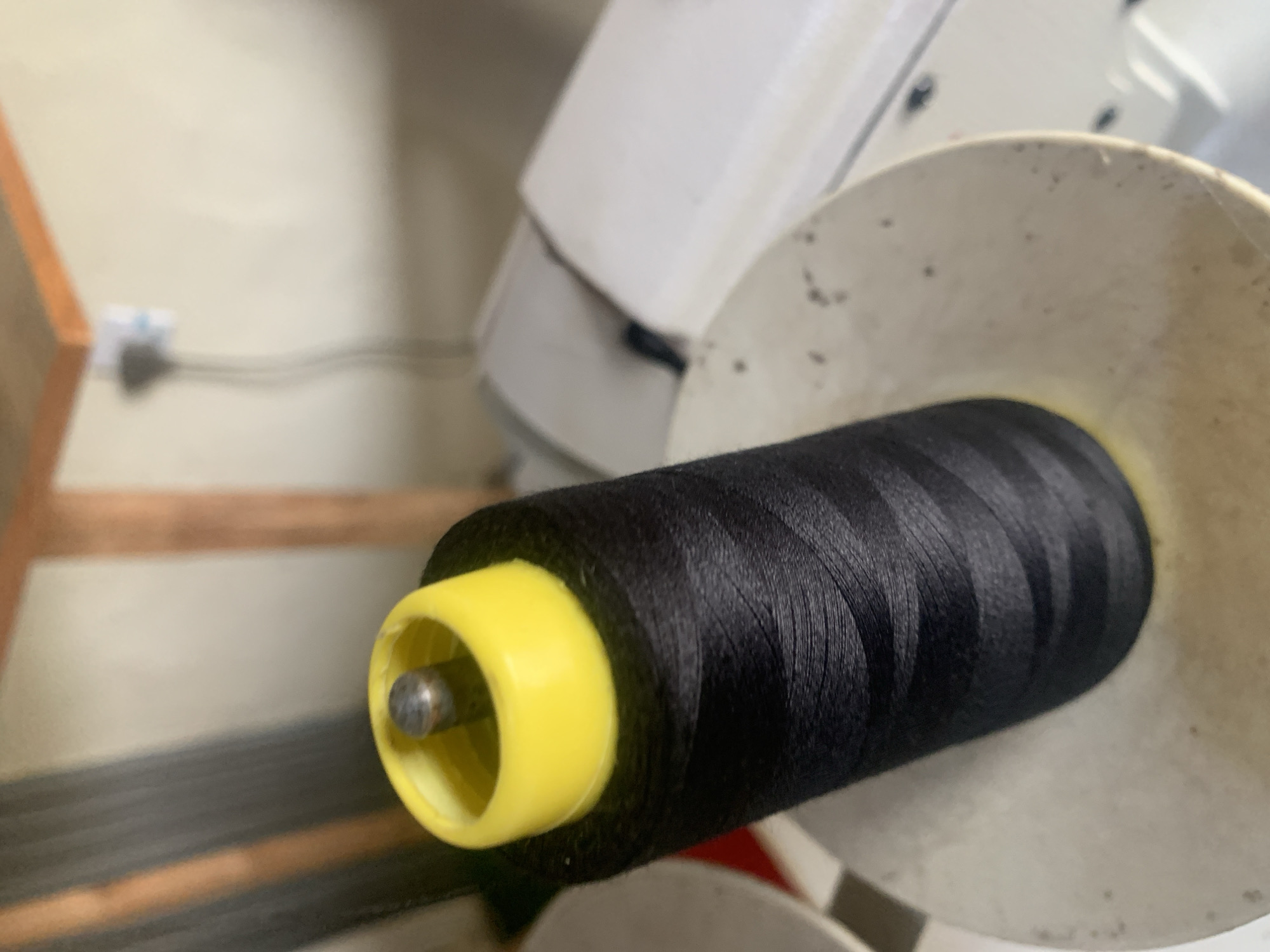 A close-up, slightly angled shot shows a spool of black thread with a bright yellow cap resting on a beige, circular platform. The thread is tightly wound and displays subtle horizontal striations. The yellow cap is cylindrical with a silver, metallic spindle protruding from its center. The background is blurred, but appears to be an indoor setting with a wall, wooden beams, and possibly industrial equipment. There are no people or animals visible in the image. The scene is well-lit, with the spool of thread being the primary focus. The location of Jauro Yinu, Nigeria is not depicted in the image.