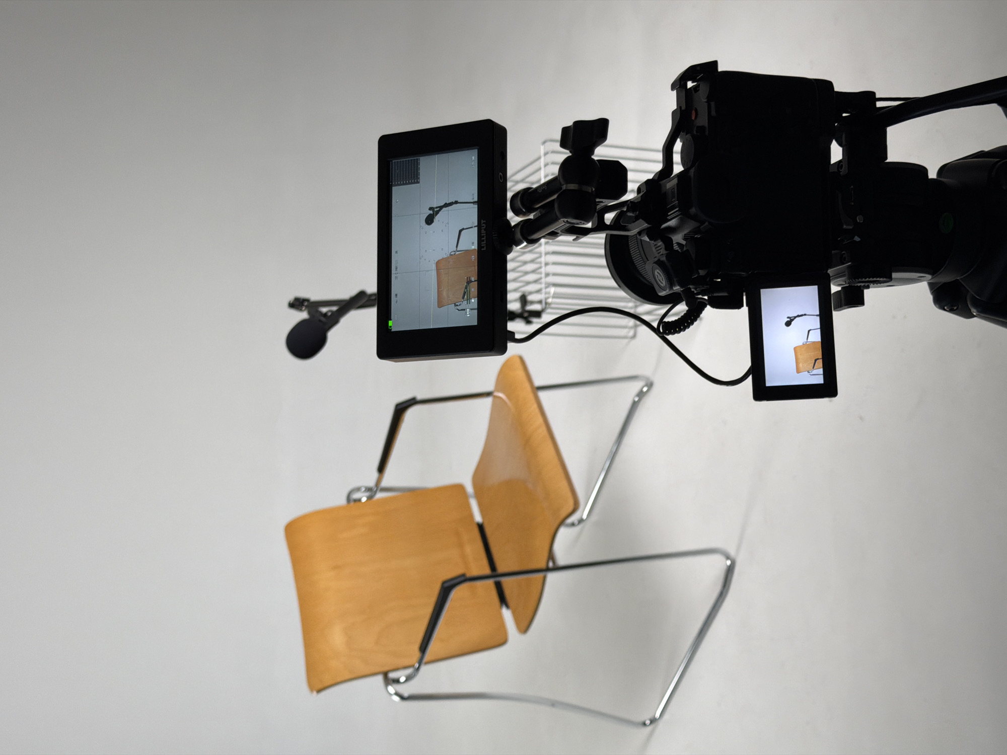 The image depicts a studio setting with a single wooden chair and two microphones positioned in front of it. A professional video camera on a tripod is aimed at the setup, with its monitor displaying a live feed of the chair and microphones. The background is a plain, light-colored wall, suggesting a controlled environment for filming or photography. There are no people visible in the scene. The location of Krasnoyarsk, Russia, is not evident from the image.