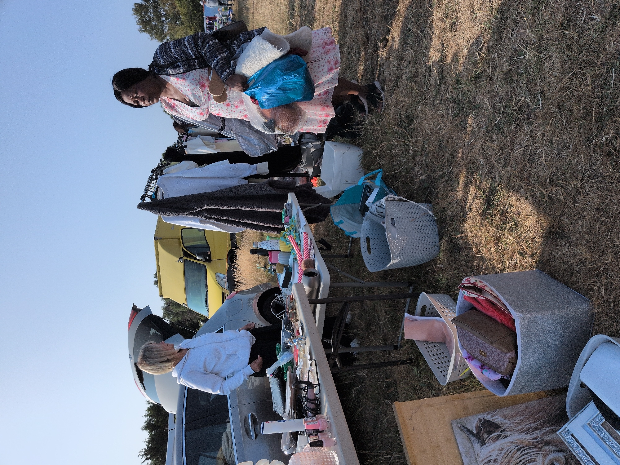 In Claverley, United Kingdom, a woman with blonde hair and a white hoodie stands near the open trunk of a car. She appears to be at a car boot sale, with tables laden with various items such as nail polish, hair dryers, sunglasses, and small decorative objects. To her right, a woman with dark hair and a floral dress is holding a blue plastic bag and some folded blankets, looking towards the tables. Clothes are displayed on a rack behind the tables. In the background, a yellow van and more cars are visible, suggesting a public event. The ground is covered in dry grass, indicating an outdoor setting.