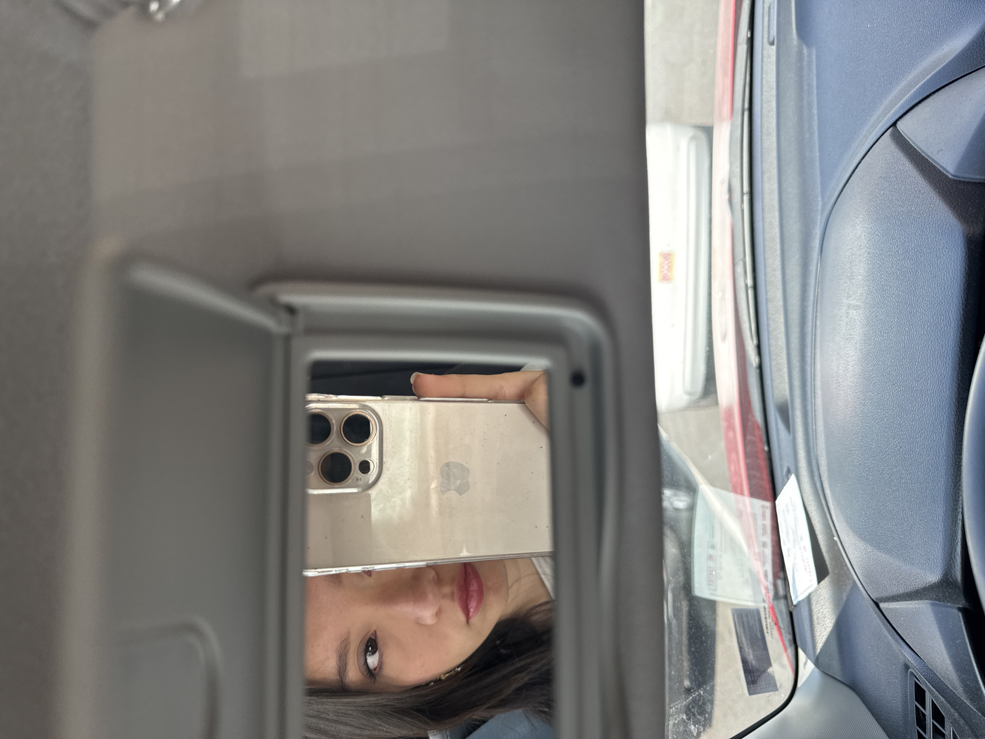 A person is reflected in the vanity mirror of a car, holding an iPhone. The camera array on the back of the phone is visible. The vehicle is parked, and the outside view shows a car and possibly a building. The location cannot be determined from the image.