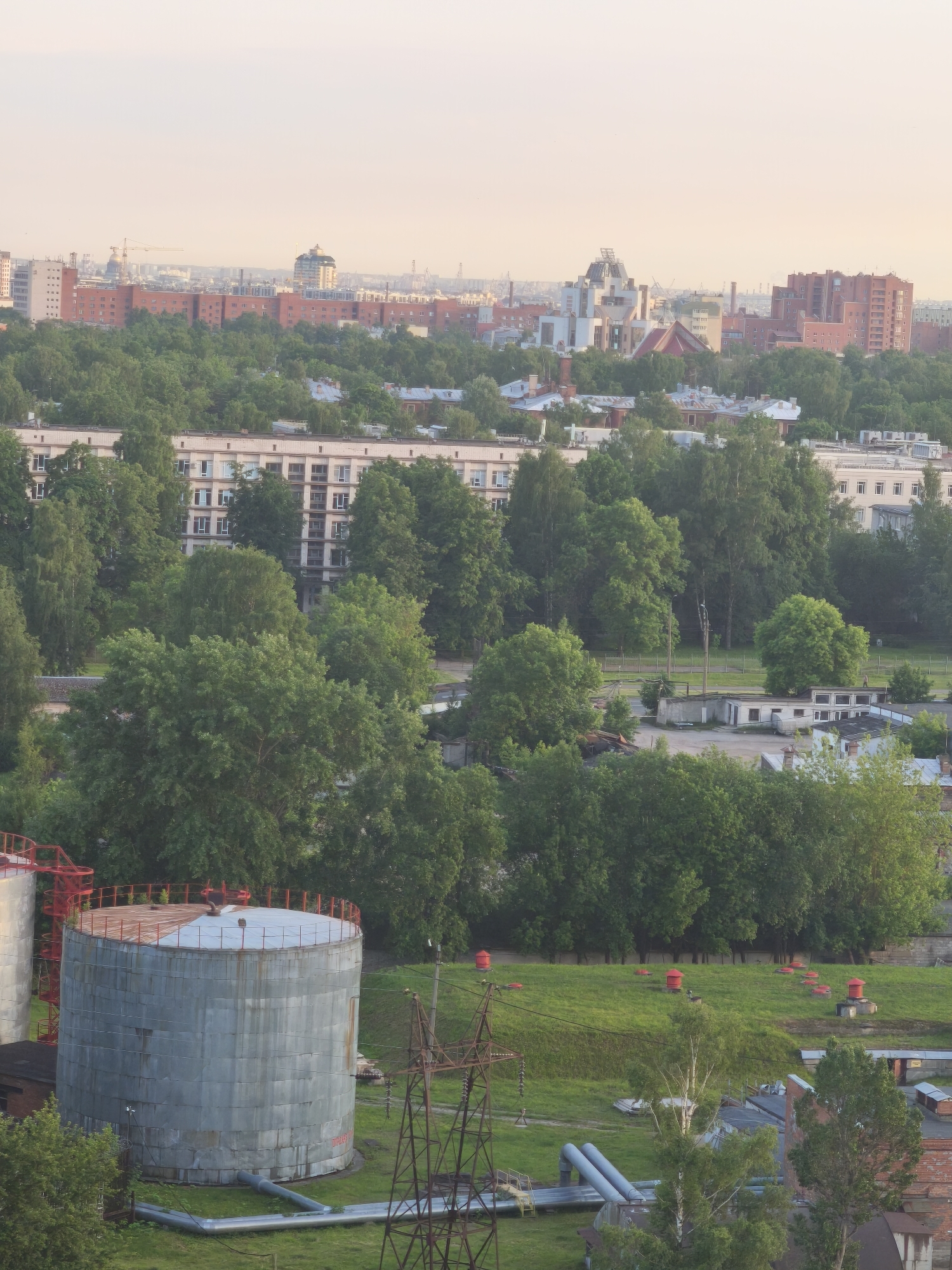 This is a high-angle, full shot photograph of an industrial area within a city, likely taken at dusk or dawn due to the soft, diffused light. In the foreground, a large, cylindrical, weathered metal storage tank with a red-railed rim is prominently featured. Adjacent to it, partially visible on the left, is another similar tank. Industrial pipes run across the grassy ground in front of the tank. A rusty metal electricity pylon stands to the right of the tank. Behind the tank, a gently sloping grassy hill is dotted with numerous small red structures, possibly vents or emergency exits, emerging from the ground. Further back, a dense canopy of lush green trees dominates the middle ground, obscuring much of the urban landscape. Beyond the trees, in the background, a mix of apartment buildings and industrial structures are visible, with some taller, modern buildings interspersed with older brick structures. The sky above is a pale, hazy orange, indicative of sunrise or sunset. The overall scene suggests an industrial zone integrated within a residential or mixed-use urban environment in Russia. There are no people visible in the frame.