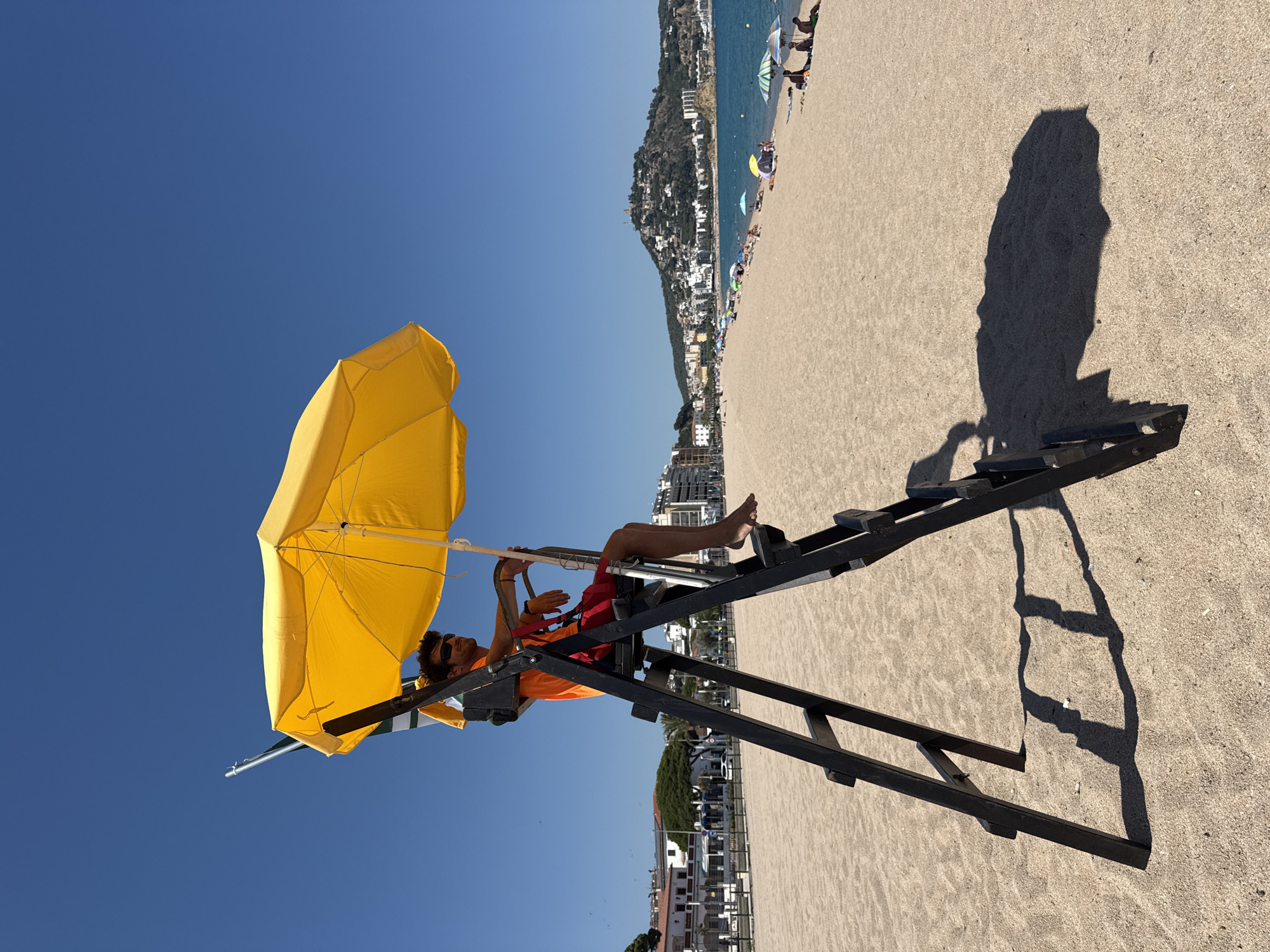 A lifeguard, wearing sunglasses, an orange t-shirt, and red shorts, sits on a high chair on a sandy beach under a yellow umbrella. The lifeguard is looking to the right. The beach is populated with several beach umbrellas and people. In the background, there are buildings, trees, and a hill with structures on it. The sea is visible to the right, with a clear blue sky above. The scene is located in Blanes, Spain.