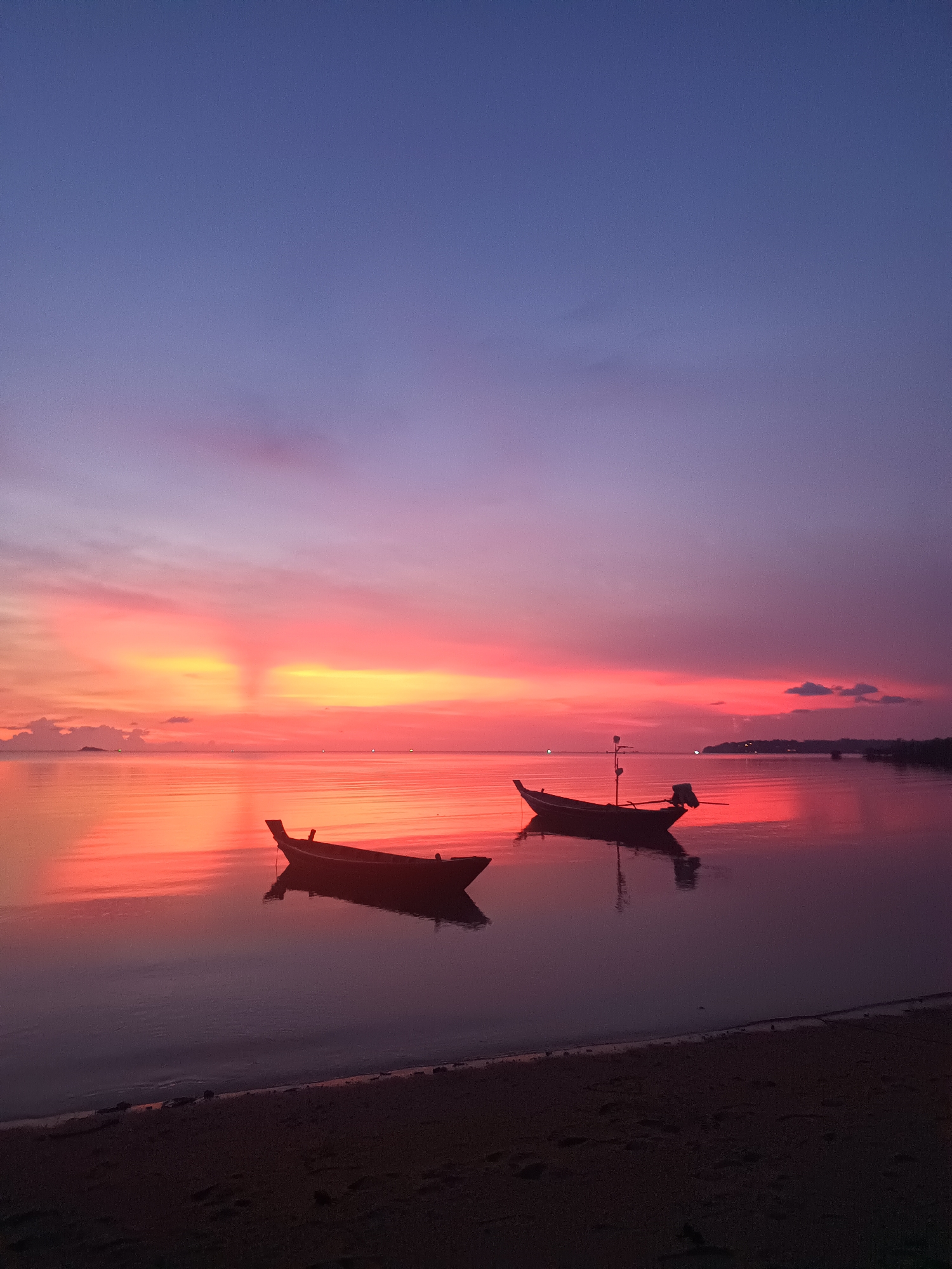 The image shows a sunset scene in Baan Wok Tum, Thailand. Two boats are silhouetted on calm water. The sky is a gradient of colors from a deep purple at the top to shades of pink, red, and orange near the horizon, with the colors reflecting on the water. The beach is in the foreground, mostly obscured in shadow. The horizon line features some landmass.