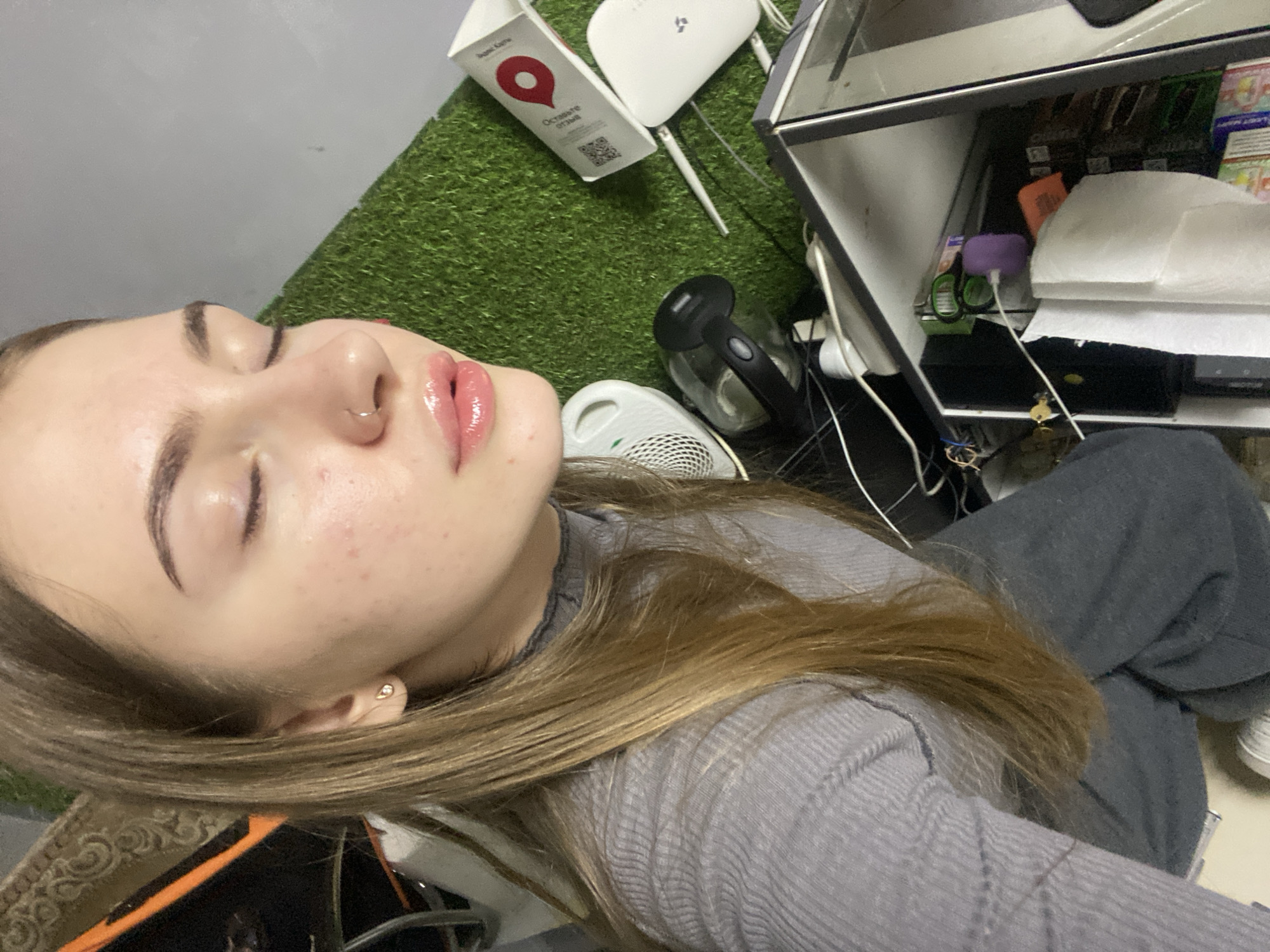 The image shows a young woman lying down, with her face close to the camera. She has light skin, dark eyebrows, and glossy pink lips. She is wearing a grey, ribbed long-sleeved shirt and dark grey pants. Her long, brown hair is spread around her head and shoulders. She has a small nose ring.
The environment appears to be indoors, with a checkered, faux-grass-like floor in the background. To the right of the woman, there is a shelf unit with various items, including what appears to be a router, a small appliance, and some papers or packaging. A white cable is visible, snaking across the scene. The lighting is bright, casting shadows. The overall impression is that of a casual, possibly domestic or business setting.