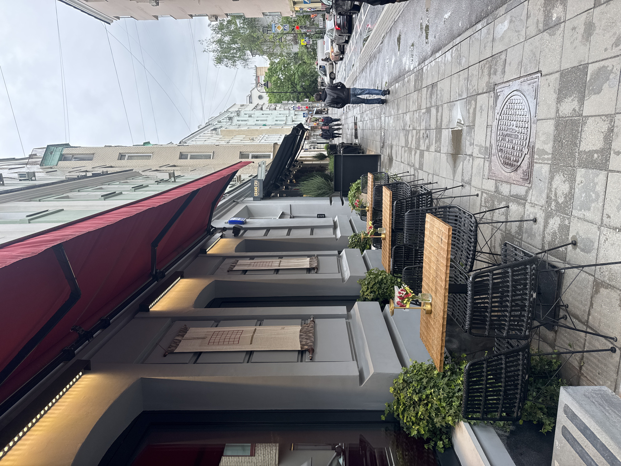 The image depicts an outdoor scene on a street in Moscow, Russia. The foreground is dominated by a building with a red awning, featuring a gray facade with architectural details like arched openings, decorative screens, and lighting. Outside the building, there are tables and chairs set up for outdoor dining, with greenery around their bases. Further down the sidewalk, pedestrians are walking, with a man in a black jacket and jeans being the most prominent. In the background, there are other buildings, trees, cars, and street signs, suggesting a busy urban environment. The street is wet, indicating recent rain or damp conditions. A manhole cover is visible on the sidewalk to the right of the seating area.