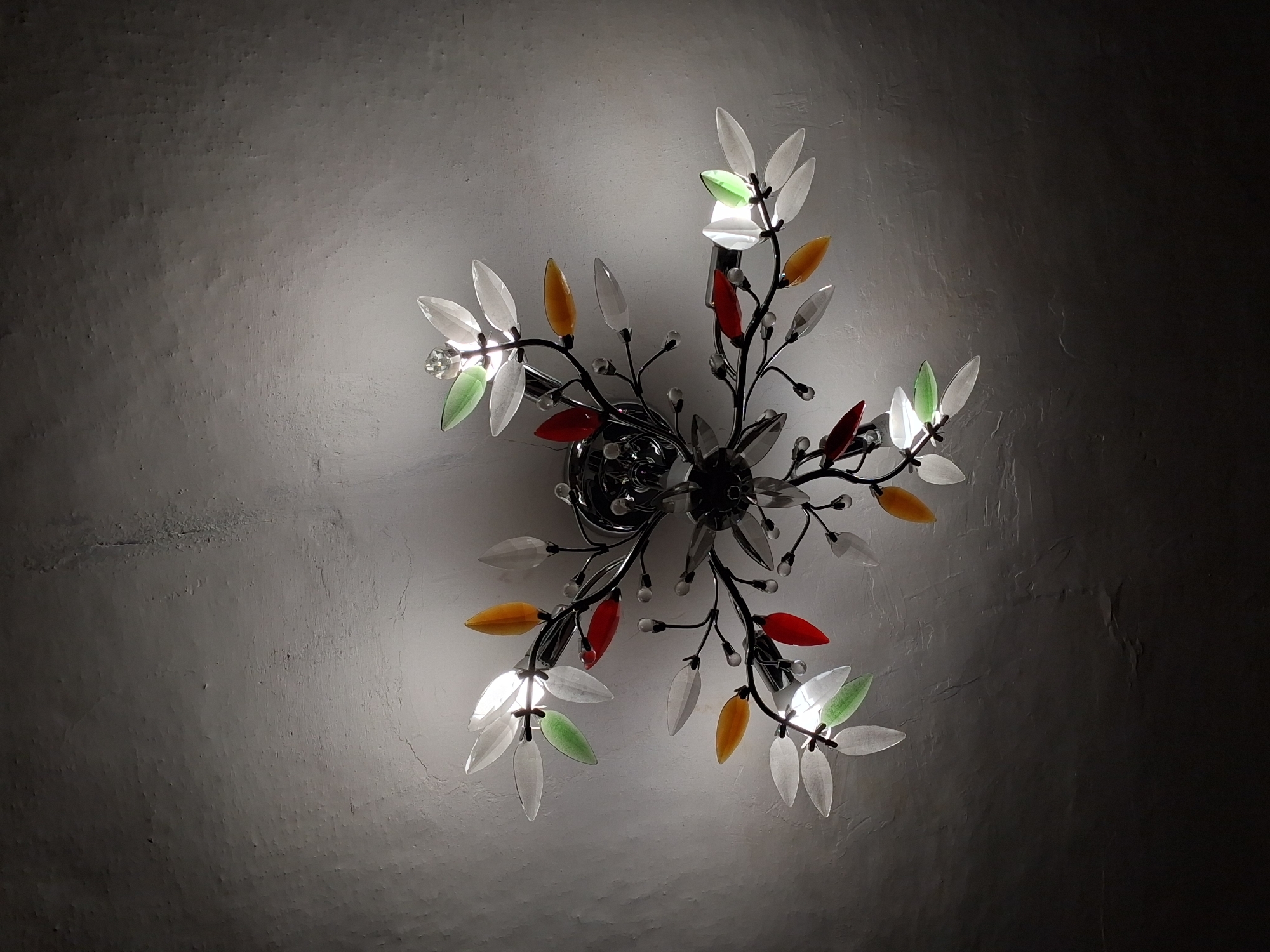 The image displays a close-up, overhead view of a decorative ceiling light fixture in Horodyshche-Kosivske, Ukraine. The fixture is designed to resemble a branching plant with several "leaves" made of clear, frosted, and colored glass or acrylic. The colors of the leaves include white, green, red, and amber. The leaves are attached to thin, dark metal branches that radiate from a central point. Small, clear, bead-like accents are also visible on the branches. The light fixture is illuminated, casting a soft glow and creating shadows on the textured, light-colored ceiling. The overall impression is one of an ornate and decorative lighting element.