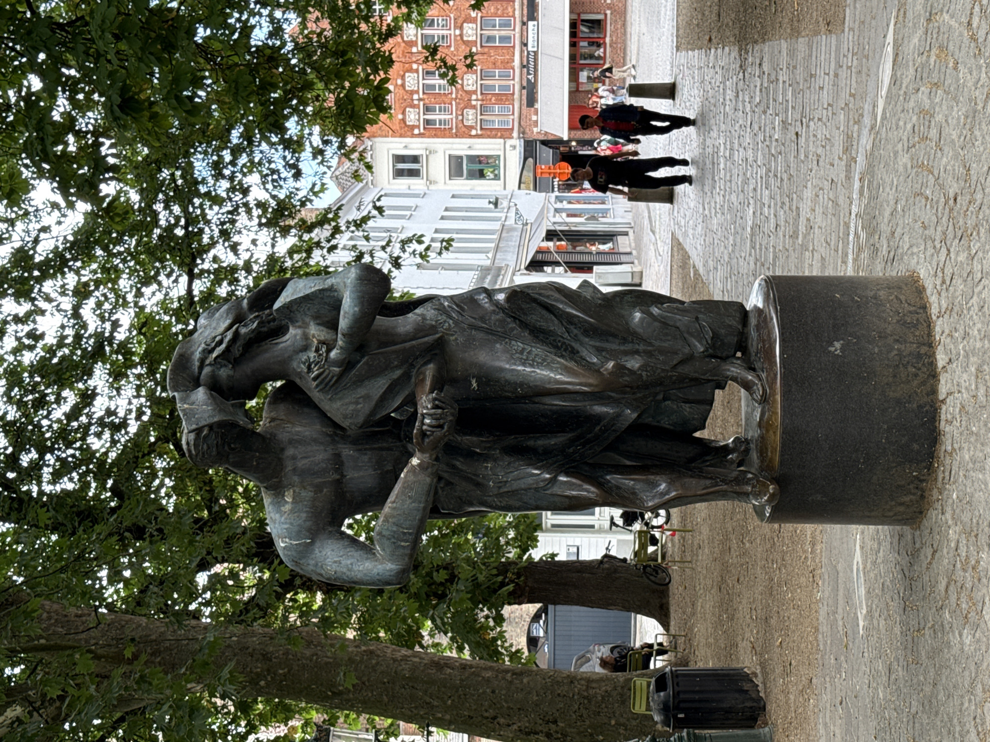 The image captures a bronze sculpture of two embracing figures in the foreground, set against a street scene in Bruges, Belgium. The sculpture, which appears to be made of weathered bronze, depicts a male figure on the left with his arm around a female figure on the right. They are both draped in flowing garments, and their heads are tilted towards each other. The sculpture is mounted on a cylindrical dark gray pedestal.
Behind the sculpture, a cobblestone path leads into the distance. On the right side of the path, two individuals are walking away from the camera. The person on the left is wearing a black t-shirt and dark pants, while the person on the right is wearing a red top and a dark jacket with dark pants. Further down the path, several other figures are visible, suggesting a public area with pedestrian traffic.
The background features a row of buildings with distinct architectural styles. The buildings are multi-story, with windows and awnings visible. Some of the buildings are painted white, while others are constructed of red brick with white accents. Trees with lush green foliage overhang the scene, providing shade and framing the sky. In the mid-ground, to the left of the path, there are two empty green chairs and a black trash bin, suggesting a seating area. A bicycle is also visible leaning against a tree in this area. The ground is covered with a mix of sand, gravel, and fallen leaves. The overall impression is that of a lively urban square or street in a European city.