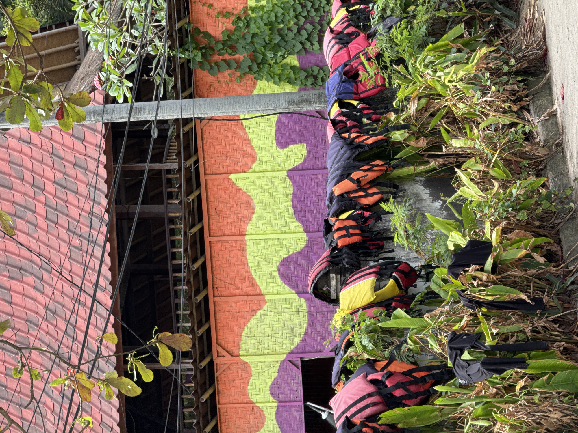 In Manggis, Indonesia, a row of colorful life vests hangs on a wall behind a lush green hedge. The life vests are predominantly red, yellow, and black, with some featuring blue accents. They are arranged in a line, some neatly folded, others slightly askew. Below the life vests, dark-colored t-shirts are draped over some of the plants. The wall itself is painted with a vibrant, wavy pattern in shades of orange, lime green, and purple. Above the wall, a red tiled roof is visible, with electrical wires crisscrossing the scene. The overall impression is one of a functional outdoor space, possibly near a body of water where water activities are offered.