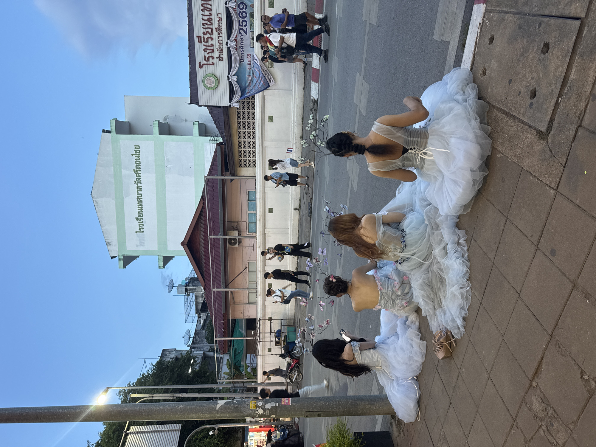 The image captures a scene on a sidewalk in Chiang Mai City Municipality, Thailand. Three young women dressed in ornate, white, tulle dresses, adorned with floral and butterfly-themed accessories, are seated on the edge of the sidewalk. Their backs are visible, revealing the intricate details of their dresses. One of the women is engaged with her mobile phone.
In the background, the city street is active with pedestrians and vehicular traffic. A white building with green accents and Thai script is visible, a sign displaying "โรงเรียนเทศบาลวัดศรีดอนไชย" is visible, indicating a school, which translates to "Municipal Wat Sri Donchai School". A banner with Thai script is also visible on the building. The sky is clear, indicating daylight. A street light is visible in the foreground.