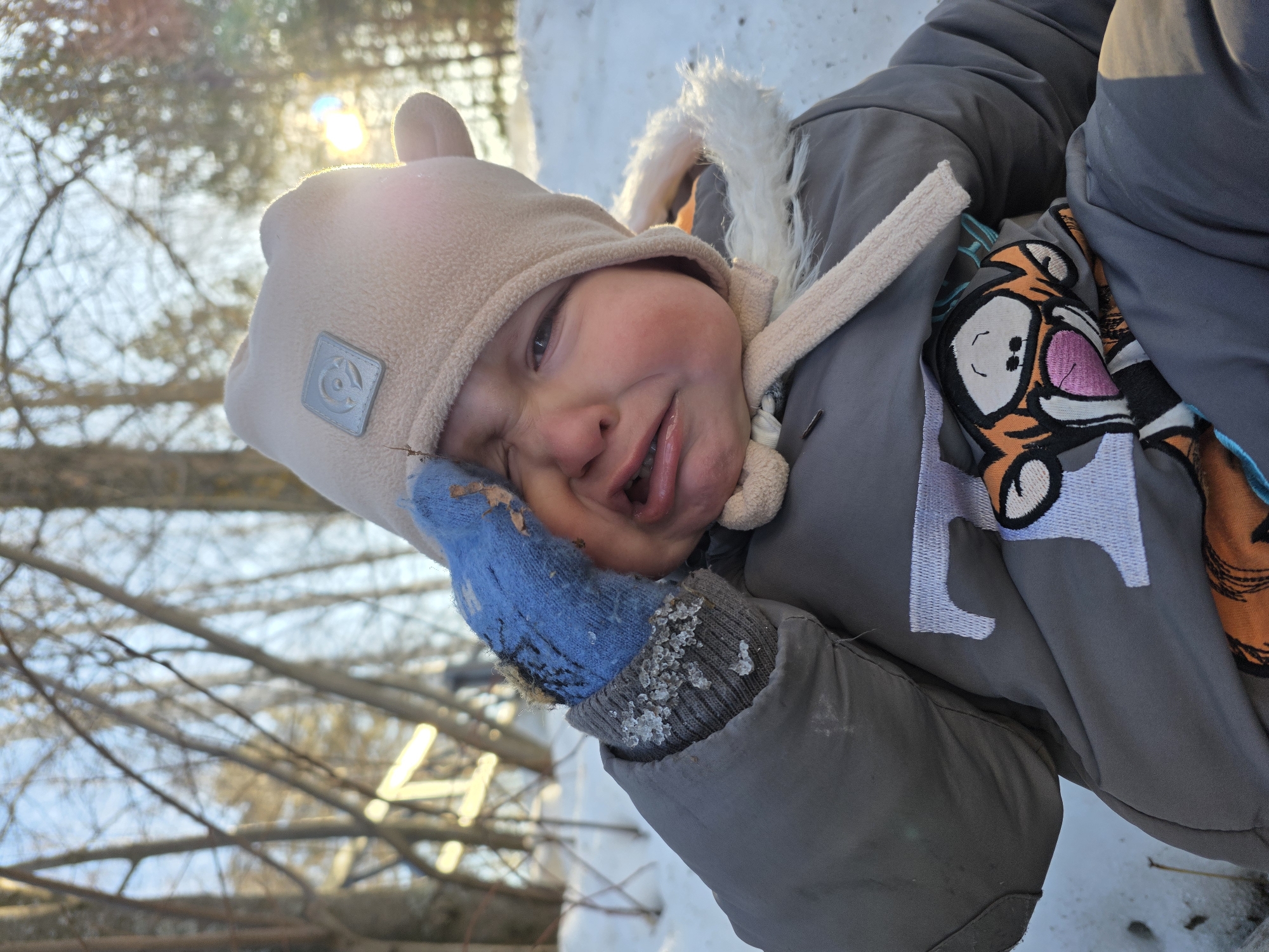 The image displays a young child seated outdoors in a snow-covered area within the city of Chaikovsky, Russia. The child, appearing to be a toddler, is exhibiting a distressed state, with a crying expression, squinted eyes, and an open mouth. The child is dressed in winter attire, including a light beige fleece hat with ear flaps and a grey square emblem on the front, and a grey insulated snowsuit featuring a white faux fur-lined hood. A colorful character patch, resembling Tigger, is visible on the chest area of the snowsuit. The child's left hand, encased in a blue knitted mitten with visible ice particles on the cuff, is raised towards their left eye area. The immediate ground is covered in white snow, with sparse bare twigs. The background consists of numerous bare trees under a bright sky, with a strong sun directly behind the child causing lens flare and backlighting.