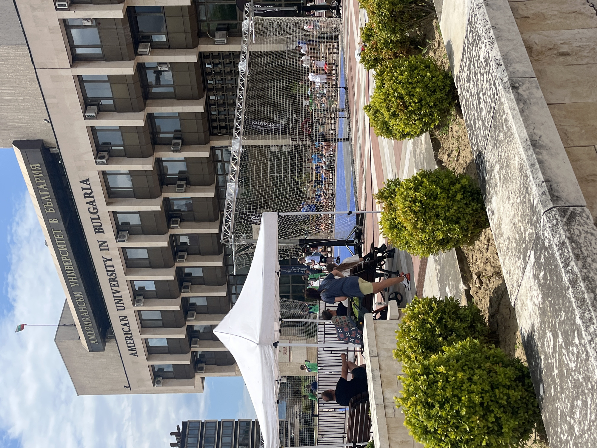 The image shows an outdoor event taking place in front of the American University in Bulgaria (AUBG) in Blagoevgrad, Bulgaria. A white tent is set up, possibly for information or registration. Several people are visible, some standing near the tent, others seated on benches. A sports net suggests a sporting event or activity is occurring, possibly a volleyball or handball match based on the court's appearance. The university building, with its prominent signage in English and Bulgarian, serves as a backdrop. Small, rounded bushes line the sidewalk. A Bulgarian flag is visible on the university building. The weather appears sunny.