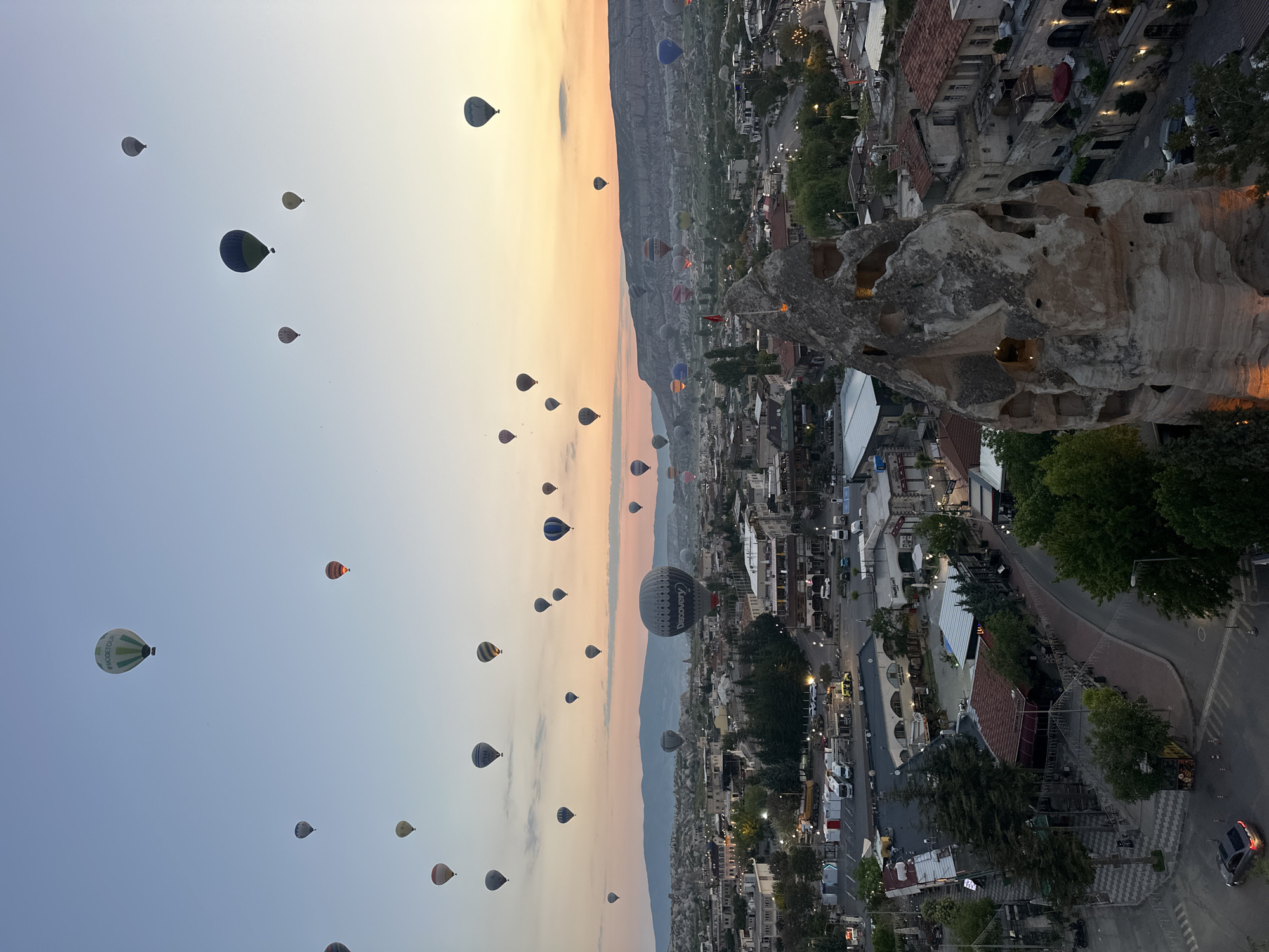 The image presents a panoramic view of Göreme, Cappadocia, Turkey, during what appears to be either sunrise or sunset. The sky, a clear canvas, transitions from a soft blue at the top to warm orange and yellow tones near the horizon, indicative of early morning or late evening light. Dozens of hot air balloons, varied in size, color, and pattern, are scattered across the sky, some ascending higher while others hover closer to the ground.
The balloons are a central subject, with specific text visible on some: "MODETOUR" is legible on a green and white balloon, and "Discovery" is prominently displayed on a large grey balloon.
Below the vibrant sky, the unique landscape of Göreme unfolds. Distinctive 'fairy chimney' rock formations, characteristic of Cappadocia, are integrated into the town's architecture. Many buildings appear to be carved directly into these geological structures, with several cave-like openings and windows illuminated, suggesting the early or late hour. A large, prominent rock formation on the right side of the frame features multiple glowing openings, seemingly part of a dwelling or establishment. The town also features roads, trees, and scattered artificial lights that are beginning to twinkle or fade. The overall scene evokes a sense of tranquility and wonder, highlighting the iconic hot air balloon experience in this historic region.