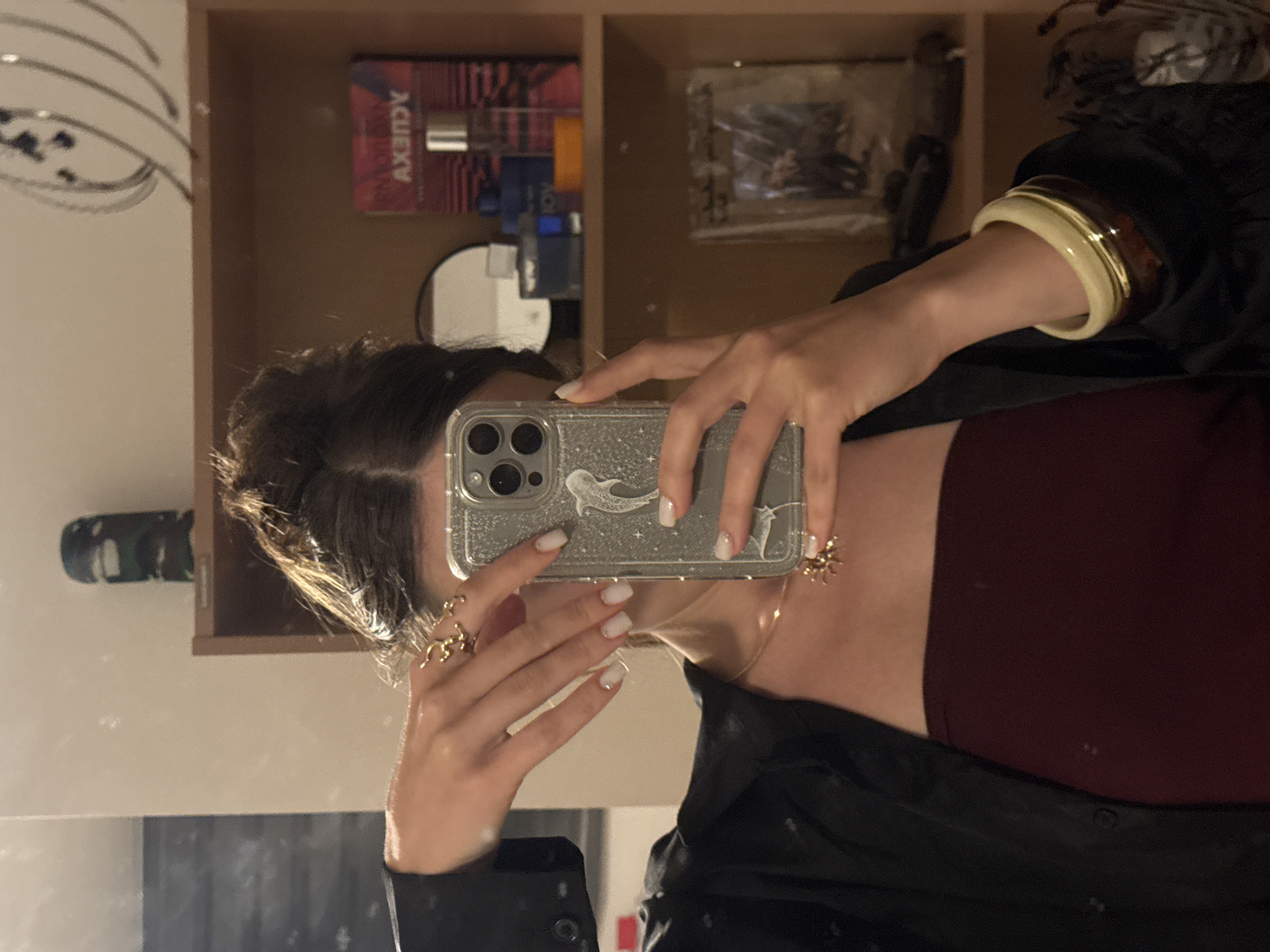 In a domestic interior setting, a person holds a silver-colored smartphone with a clear, glittery case in their right hand, angled to capture their reflection in a mirror. The phone's case features a silver dolphin sticker. The person's left hand is positioned near their face, with fingers adorned with white nail polish and several gold rings, including a distinctive abstract design on the index finger. They are wearing a dark, possibly black, blazer over a deep red, strapless top. A delicate gold chain necklace with a spider pendant is visible around their neck. On their left wrist, they wear two stacked bangles, one appearing to be a creamy white or beige and the other a darker, possibly amber or brown, material. The background reveals a wooden shelf unit. On the shelves, various items are visible, including a cylindrical dark object with a camouflage pattern, perfume bottles, a framed picture, and decorative metallic ornaments hanging from above. The setting does not contain any identifiable elements to place it specifically in Budva, Montenegro; it depicts a generic indoor environment.