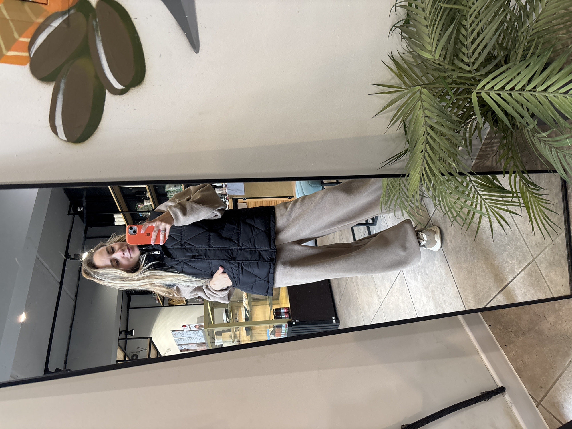 An indoor, full-body mirror selfie features a Caucasian female in what appears to be a cafe or similar commercial establishment in Chaikovsky, Russia. The woman has long, straight blonde hair styled in a high ponytail and is looking at the screen of an orange-cased smartphone held in both hands, suggesting she is capturing the image. She exhibits a subtle smile.
She is dressed in a black, quilted, collared vest worn over a beige, long-sleeved top. Her lower attire consists of wide-leg, beige athletic pants or sweatpants. On her feet are light-colored sneakers with white soles and beige/grey uppers. Her left hand is partially inserted into the pocket of her vest, while her right thumb is positioned near the side button of the smartphone.
The woman and the background are reflected in a large, rectangular mirror framed in black, positioned on the left side of the frame. The reflection reveals a modern interior with a light grey ceiling featuring exposed ductwork and black track lighting fixtures. To the left of the woman's reflection is a glass display case with multiple shelves, containing various food items or products. Further back and to her right, black open shelving units hold bottles, containers, and mugs. A light blue upholstered stool is partially visible behind her lower legs. Wall-mounted menus or posters are also discernible in the reflection.
To the right of the mirror, the wall is painted an off-white or light beige color. A mural on this wall depicts three brown, oval-shaped coffee beans with white horizontal highlights, positioned above a dark grey triangular shape, and partially visible orange elements suggesting stylized packaging or branding. The floor throughout the visible area is composed of light-colored square tiles with dark grey grout lines, laid diagonally. In the lower right foreground, a dense, green artificial plant with palm-like fronds partially obscures the wall and floor. A black corrugated pipe is affixed vertically to the white baseboard on the wall to the immediate left of the mirror's frame.