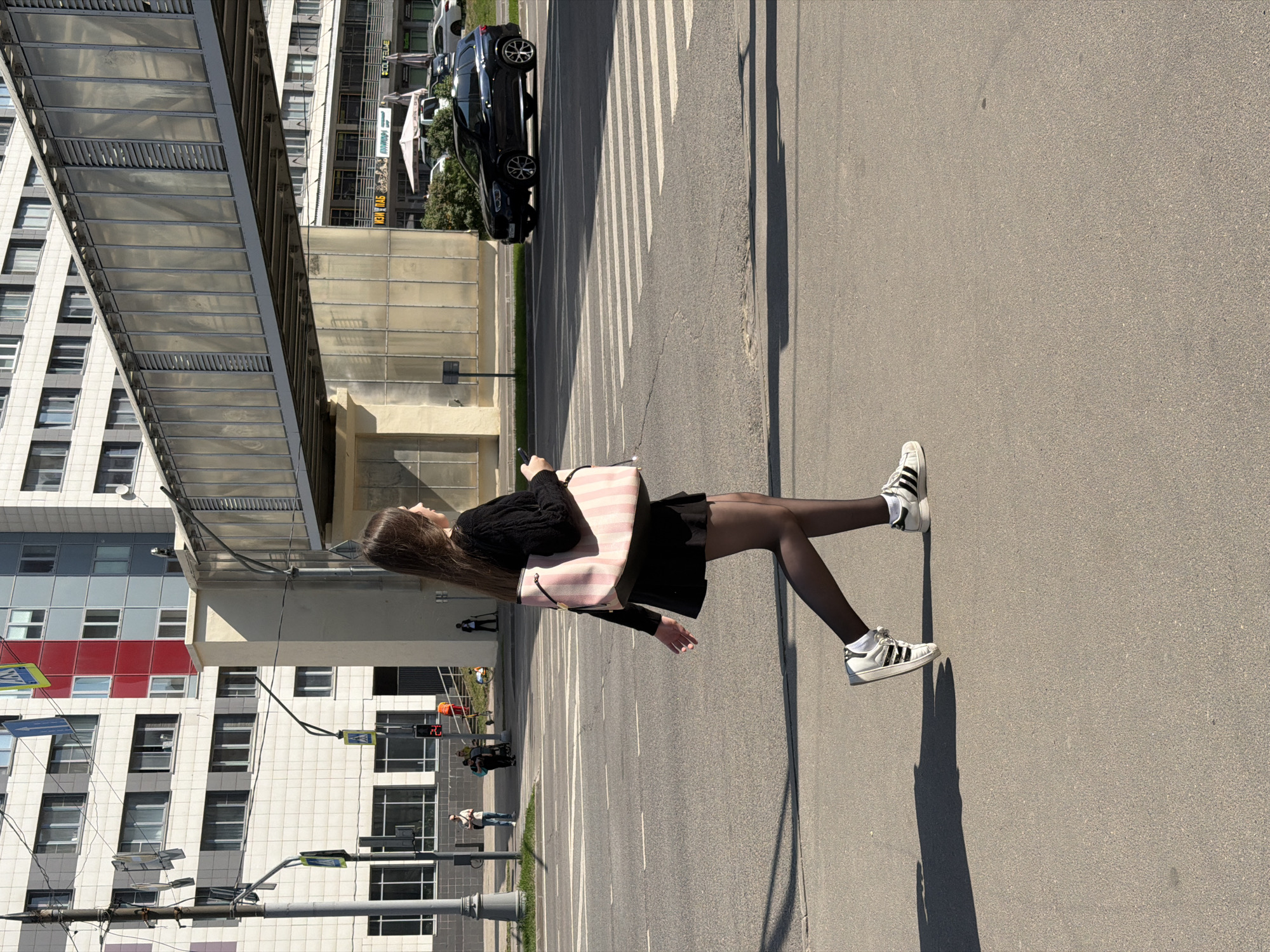 A woman in a black sweater, black skirt, black tights, and white sneakers with black stripes walks across a crosswalk on an asphalt street. She carries a large pink and white striped tote bag. The image is shot from a low angle, looking up at the woman. The background features a building with many windows and a pedestrian bridge. The location is Moscow, Russia. Cars are visible in the background.
