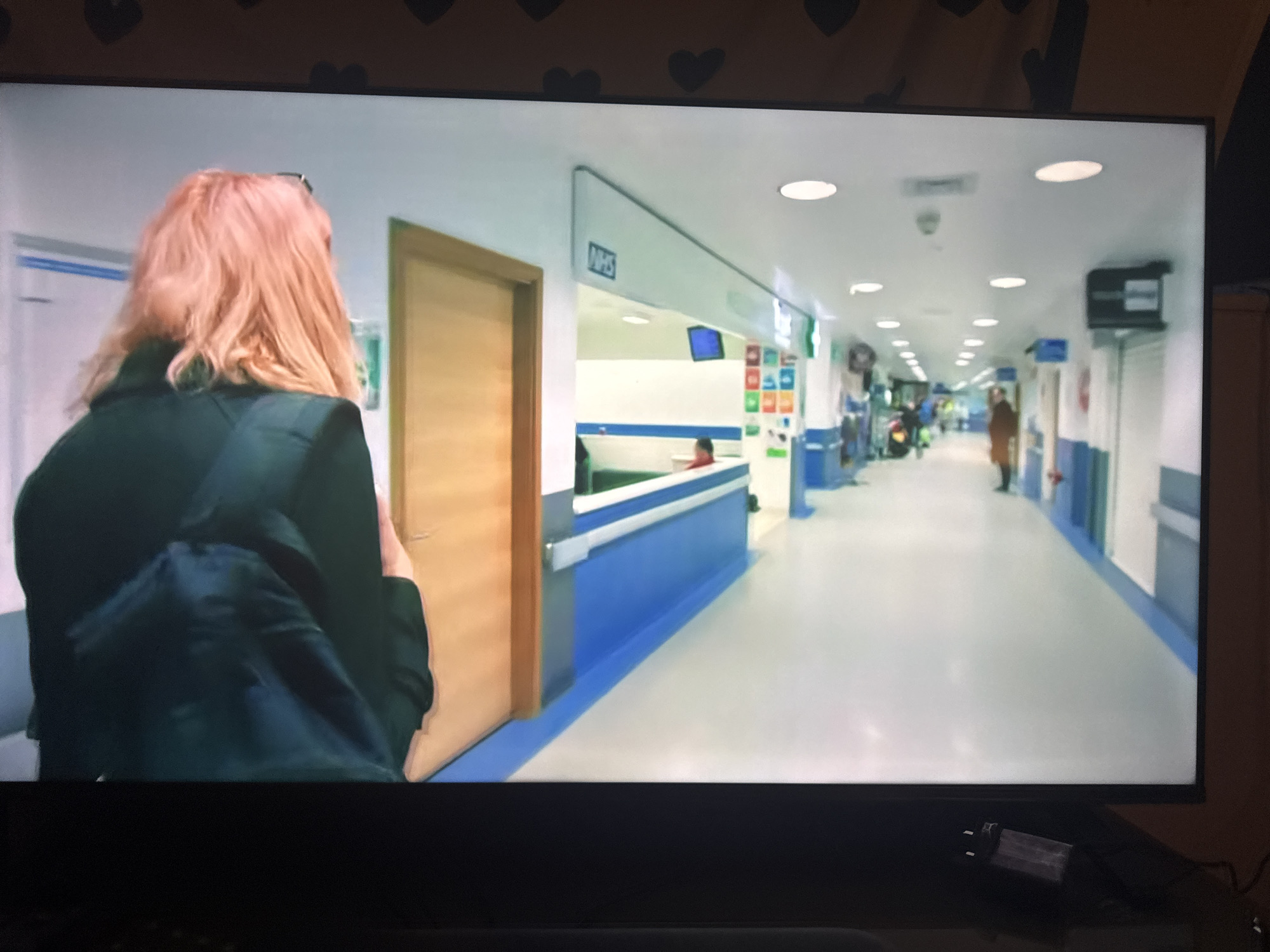 The image shows a television screen displaying a scene from what appears to be a hospital corridor. A person with reddish-blonde hair, wearing a dark jacket and backpack, is standing in the foreground, facing away from the camera and towards the corridor. The corridor itself is long and brightly lit, with white walls and blue accents along the lower section. There are doors on either side, and signs, including one with "NHS" visible. In the distance, several other individuals can be seen walking or standing. The scene is generic and does not provide any specific visual cues to suggest it is located in the city of Cheltenham, United Kingdom.