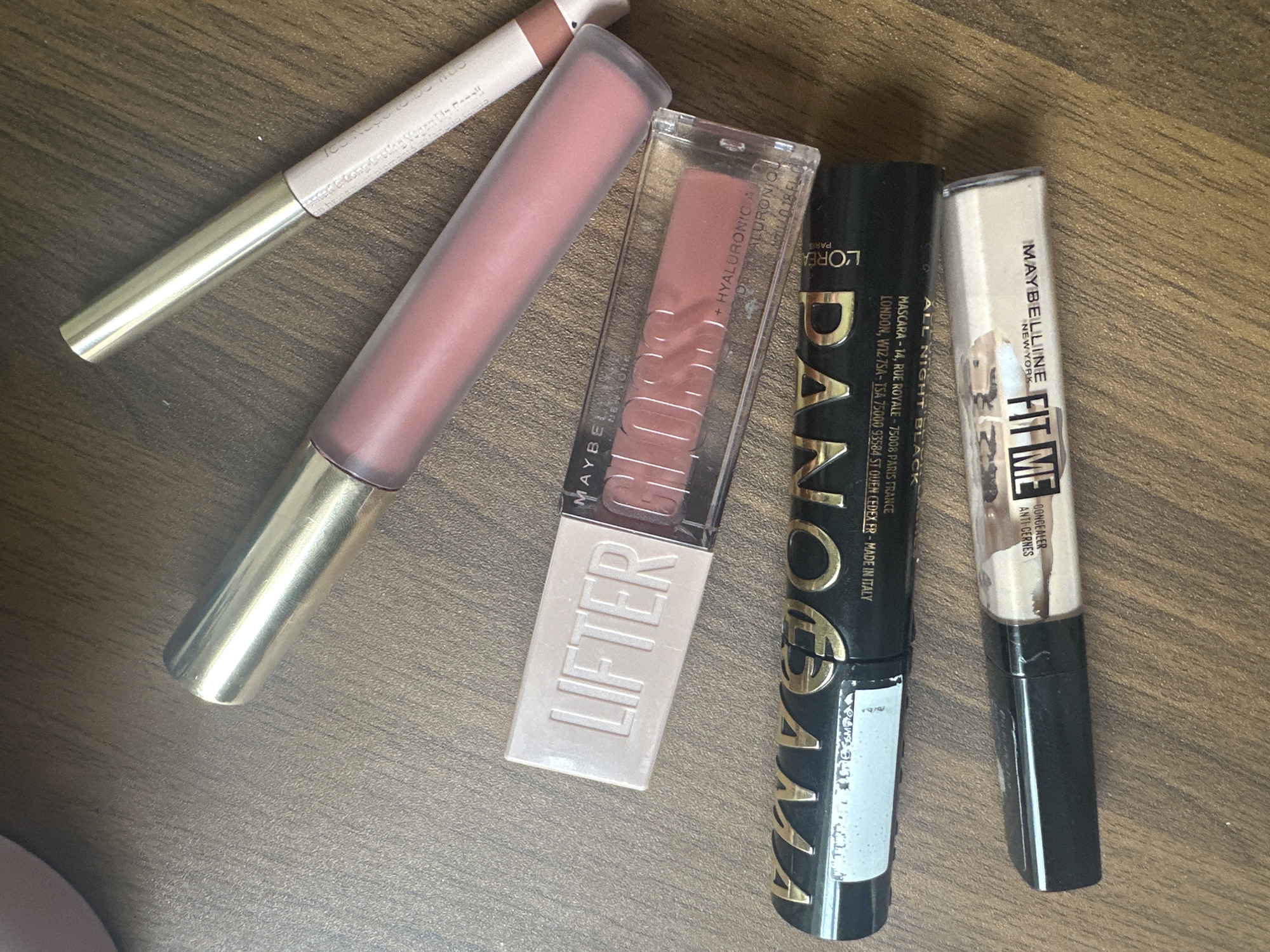 The image shows a collection of makeup products arranged on a wooden surface. From left to right, there is a lip liner pencil, a pink lip gloss with a gold applicator, Maybelline Lifter Gloss, L'Oreal Panorama All Night Black mascara, and Maybelline Fit Me concealer. The location is not discernible from the image.