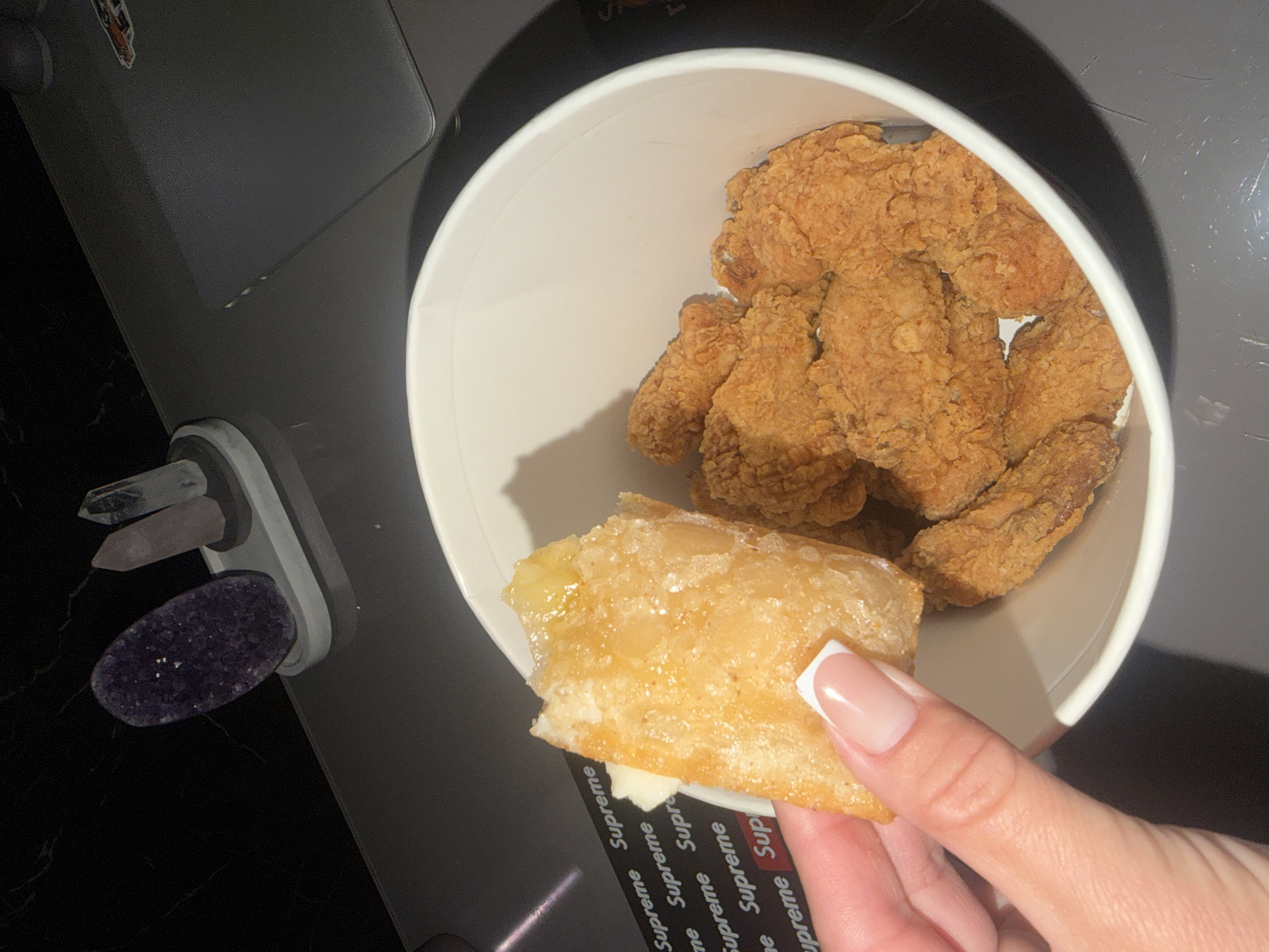 A hand with a French manicure holds a piece of fried food, possibly a spring roll or egg roll, that is coated in a golden, glistening glaze, over a white bucket filled with fried chicken pieces. The background shows a dark, reflective surface, potentially a desk, with some decorative crystals and a laptop. The overall scene appears to be indoors, on a flat surface, with artificial lighting. There is no identifiable information suggesting the location is in Novosibirsk, Russia.