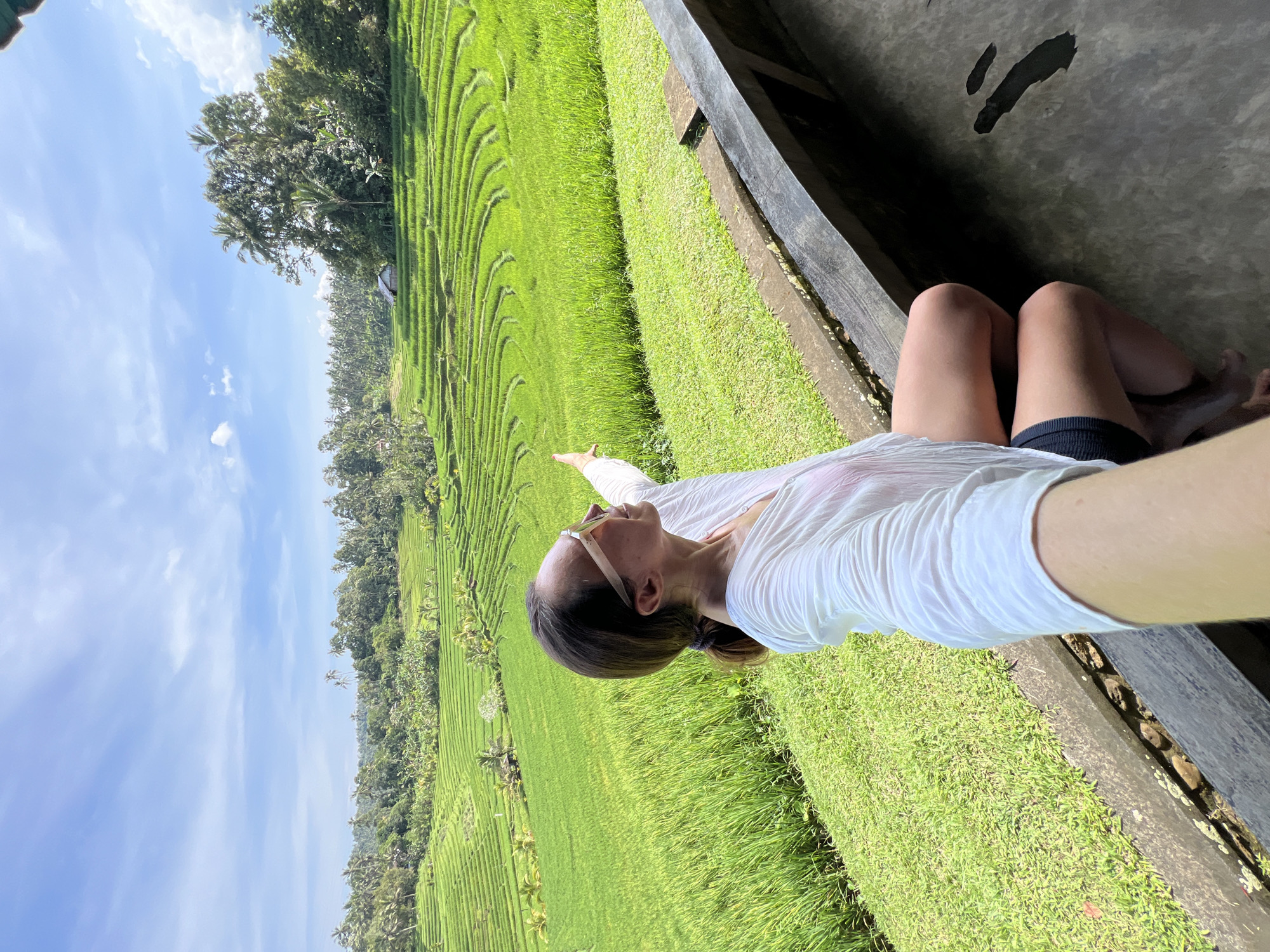 A person is taking a selfie in a rural setting. The individual is wearing a white top, dark shorts, and sunglasses, lying down and extending their arm to take the photograph. The background features a vibrant green rice field with terraced patterns, interspersed with trees and a clear blue sky with scattered clouds. The scene is likely located in Pupuan, Indonesia, known for its rice terraces.