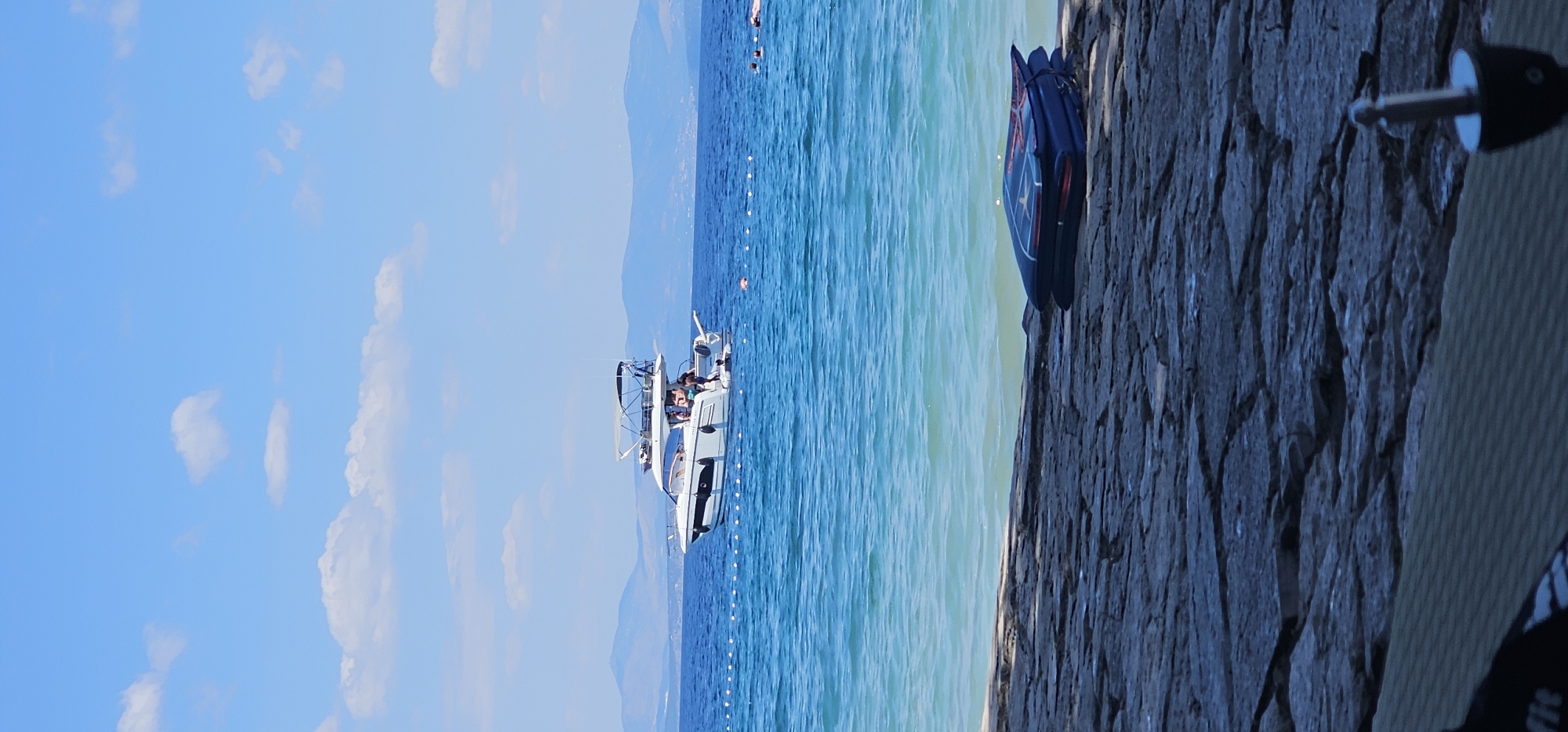 The image depicts a serene coastal scene in Supetar, Croatia. The turquoise water of the Adriatic Sea is calm, with gentle waves lapping at the rocky shore in the foreground. A white motor yacht with a hardtop is anchored offshore, carrying several individuals who appear to be enjoying a day on the water. A series of white buoys mark a perimeter in the sea. In the background, the hazy silhouette of distant hills suggests the presence of mainland or other islands. The sky is a clear blue, dotted with a few fluffy white clouds. On the rocky shore, a folded blue mat with red accents rests near the water's edge. The overall impression is one of a tranquil summer day at the beach.
