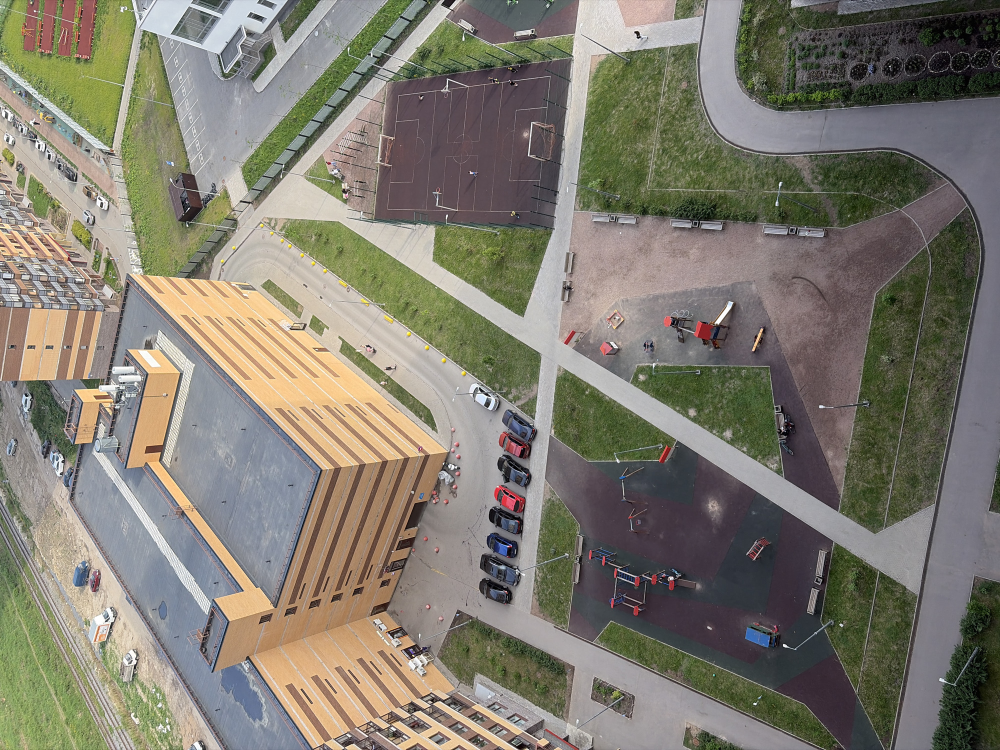 The image presents a high-angle view of a residential area in what is likely Saint Petersburg, Russia, given architectural styles and vegetation. The perspective is looking down, showing rooftops, roads, playgrounds, and recreational spaces.
Prominent is a multi-story building with a contrasting striped facade of beige and brown vertical panels. The roof of the building is dark gray, with structures presumably housing ventilation or other mechanical equipment.
Alongside the building, several cars are parked along a paved road. These cars include a mix of dark and red vehicles, appearing to be primarily passenger cars. Further down the road, there are a few parked vans or trucks.
Recreational spaces visible include a multi-purpose sports field, a playground with colorful equipment (red, blue, gray) and surfaces in various colors including a brownish purple surface, a sandy area with playground equipment, and a paved area with benches. Sidewalks and paths connect these areas and cross through grassy plots.
The road network consists of gray asphalt surfaces with white lines marking parking spaces. Yellow bollards run along the road. Vegetation includes lawns, trees, and bushes.
There are a small number of individuals scattered throughout the image, engaging in activities, such as walking.