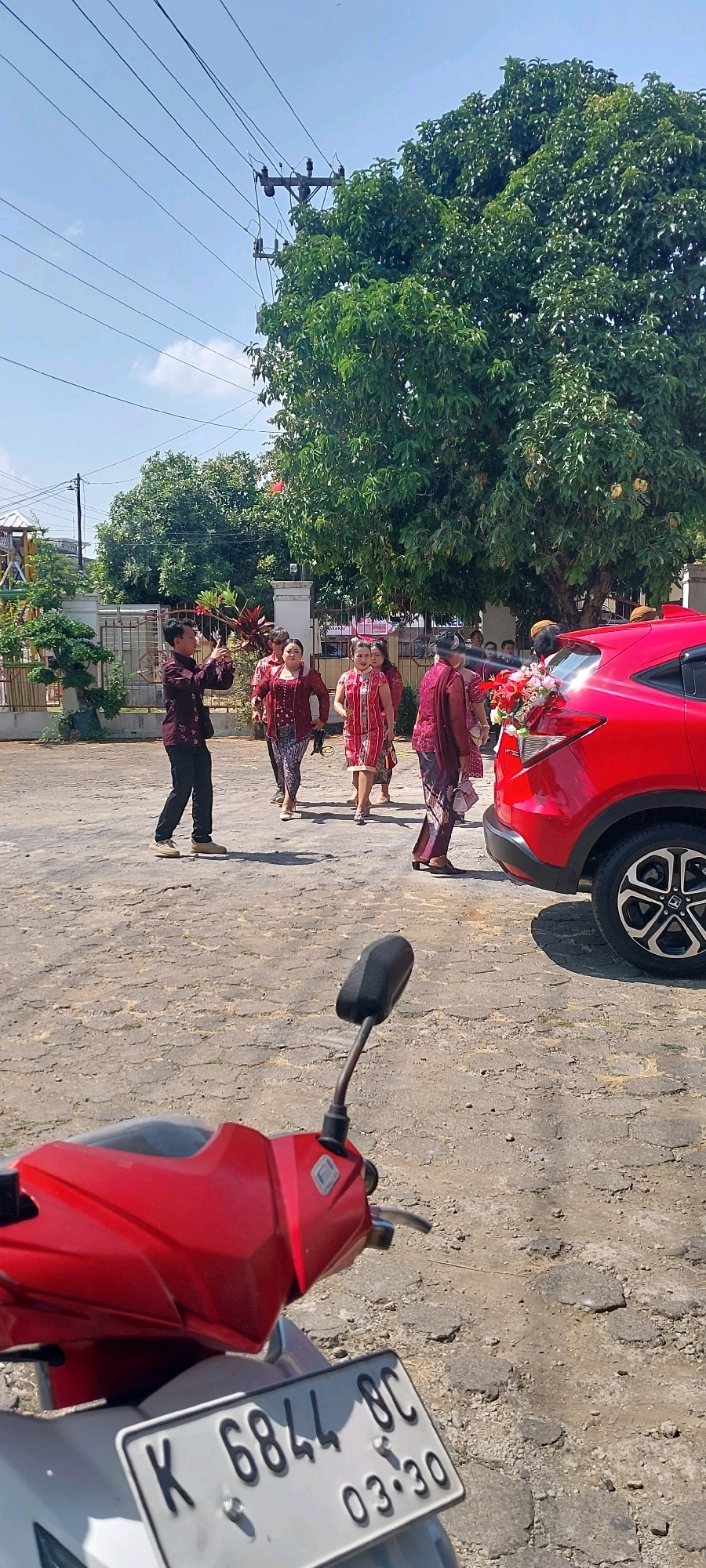 In Keling, Indonesia, on a paved outdoor area under a clear sky with scattered clouds, a group of people are gathered. A man in a dark red patterned shirt and black pants is taking a photo with his phone, facing a procession of women in traditional red and white patterned attire walking towards a red car. The car is decorated with a floral arrangement on the back. Another red motorcycle is visible in the foreground, its license plate reading "K 6844 BC 03.30". Trees and a building with a white fence are in the background.