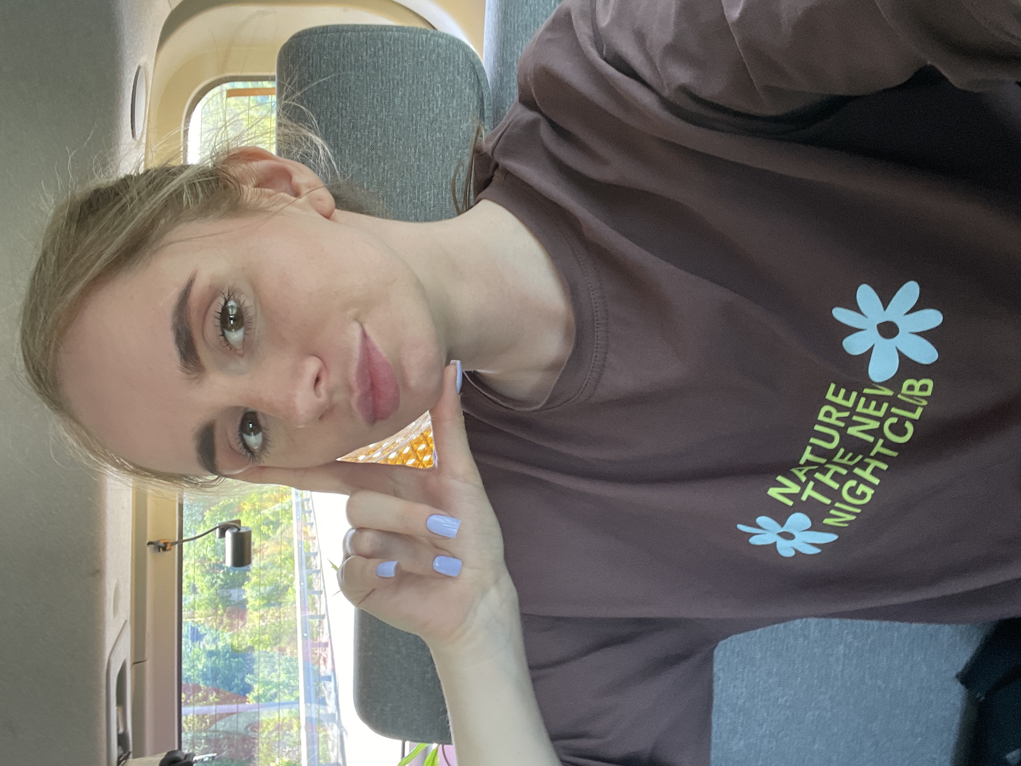 A young woman is taking a selfie while sitting inside a vehicle, likely a car or van. She is light-skinned with light brown or blonde hair pulled back. She is wearing a brown t-shirt with light blue and yellow graphic text and flower designs on the right side. Her fingernails are painted a light purple color. The vehicle's interior is light-colored. Through the window, trees and possibly other foliage can be seen, along with what appears to be an urban setting in the distance. There is nothing to indicate this image was taken in Vladivostok, Russia.