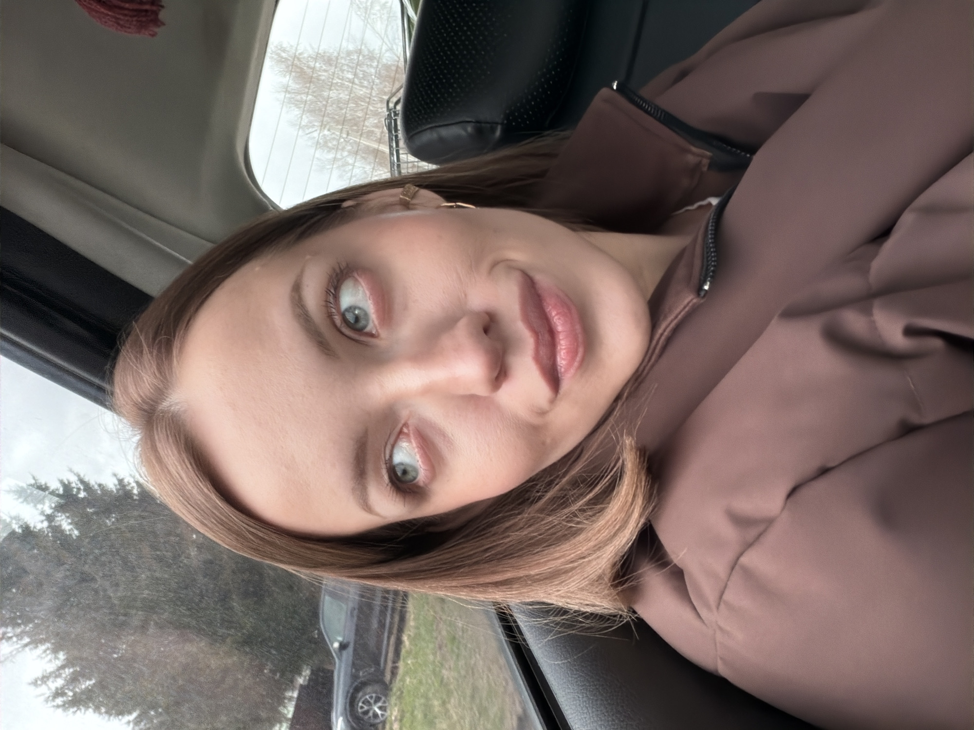 A selfie-style portrait shows a young woman inside a car, looking directly at the camera with a wide-eyed expression and a soft smile. Her head is tilted, and her body is oriented as if in the passenger seat. She has medium-length, light brown hair, and appears to be wearing light makeup, including mascara and a glossy pink or peach lip product. A small gold earring is visible on her right earlobe.
She is dressed in a brown or mauve zippered jacket, worn over what appears to be a white garment, visible at the partially unzipped neckline. The jacket has a slightly puffy, insulated appearance. The car's interior is visible, including part of a dark headrest behind her and the car's ceiling lining.
Through the car windows, an overcast sky and the branches of deciduous trees, some bare, suggest a late autumn, winter, or early spring setting. To the left, a dark-colored car is partially visible in the distance, parked or driving on what appears to be a rural or semi-rural road with visible ground. The overall impression is daytime with muted light due to the cloud cover. There is no visible text in the image. The location context for this media is Раздолье, Russia.