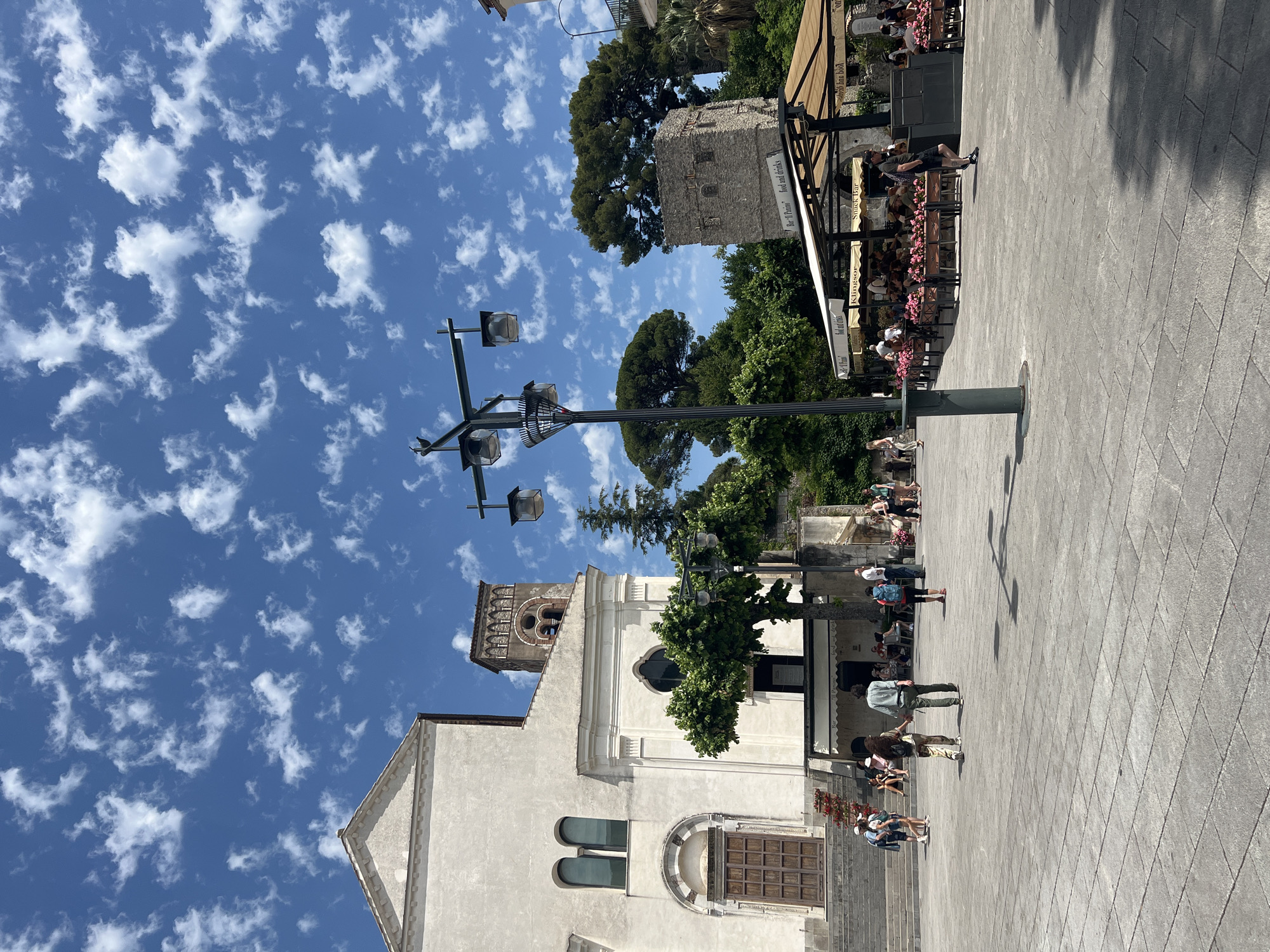 A sunny day in Ravello, Italy, features a paved piazza with a church and a cafe. The church, on the left, has a beige facade, tall arched windows, and a wooden double door. A bell tower with arched openings rises behind it. To the right, a cafe with beige awnings and outdoor seating is visible, with people seated at tables. A stone tower, possibly an ancient remnant, stands behind the cafe. A lamppost with multiple lamps stands in the center of the piazza. Scattered clouds dot the bright blue sky. Several people are walking in the piazza and near the cafe.