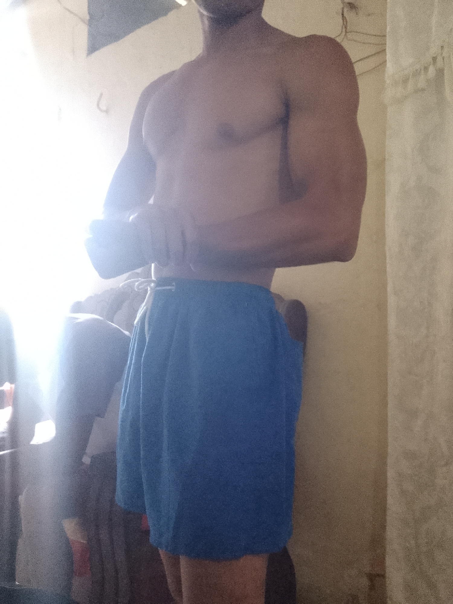 A shirtless man in blue shorts stands in a room with cream-colored walls. A window with bright light is to the left of the man. The exact location in Juan Griego, Venezuela, is not discernible from the image.
