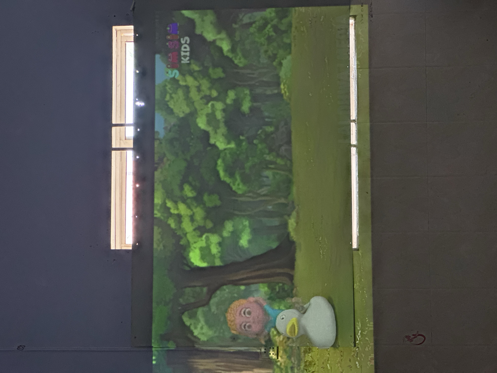 The image shows a projected scene of a cartoon character, a child sitting on a white duck, in a forest environment with trees. The logo "SimSim KIDS" is visible in the upper right corner of the projected image. Below the projected image, the wall appears to be tiled, with a red marking that resembles a stylized symbol on the lower left. Two windows with bright light coming through them are visible above the projected image. This scene is likely in a classroom or a room set up for children's activities in Kampot, Cambodia.
