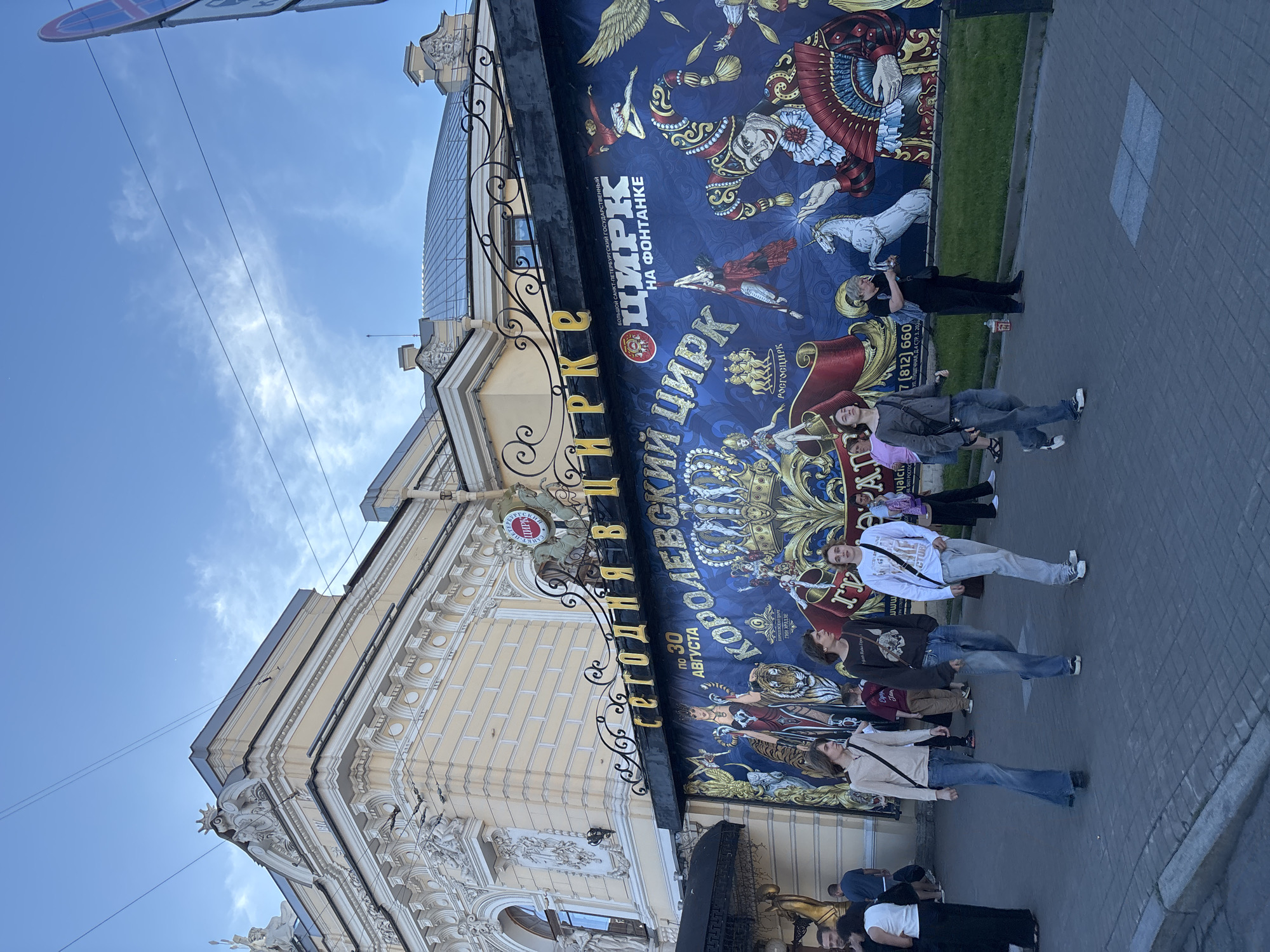 The image depicts the Bolshoi Saint-Petersburg State Circus on Fontanka, a well-known circus in Saint Petersburg, Russia. The facade of the circus is adorned with a large, vibrant mural featuring circus performers, animals, and ornate decorations. The text on the mural, "СЕГОДНЯ В КОРОЛЕВСКИЙ ЦИРК НА ФОНТАНКЕ" translates to "TODAY IN THE ROYAL CIRCUS ON FONTANKA". The building itself is an older, ornate structure with classical architectural elements. In front of the circus, a group of young people are walking on a cobblestone sidewalk. The sky above is blue with scattered clouds, and power lines are visible crisscrossing the sky. The overall scene captures a moment in front of a historic cultural landmark on a day with pleasant weather.