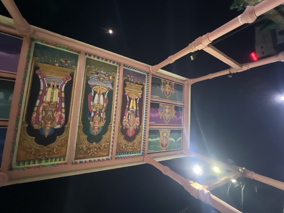 The image displays an ornate, light-colored (possibly pink or peach) overhead structure against a dark night sky. This structure is composed of sturdy beams or pipes forming a rectangular grid, likely part of a canopy or architectural installation.
Embedded within the framework are multiple rectangular art panels. The most prominent panels on the left feature vibrant, detailed depictions of figures, likely Hindu deities, standing upside down on ornate architectural bases. Their heads point downwards within the panels, and they are richly adorned in traditional style. To the right, a column of three smaller panels continues this theme, displaying similar inverted figures or reflections of architectural elements against different colored backdrops (purple, green).
The background is a clear night sky, with a bright, distinct moon visible in the upper left. Several intense artificial light sources, likely spotlights, illuminate parts of the structure from below and to the right, creating bright glares.
On an upper beam of the structure, in the top right quadrant, red illuminated text is visible, appearing upside down from the camera's perspective. The text clearly reads "SHOOKS" or "SKOOKS". The overall presentation, particularly the inverted figures and text, suggests an intentional artistic or architectural design, possibly involving reflections or a specific viewing experience common in certain temple settings. Given the Palani location context, this is likely an adorned outdoor element of a temple or religious site. The scene is static, capturing the illuminated structure against the nocturnal expanse.