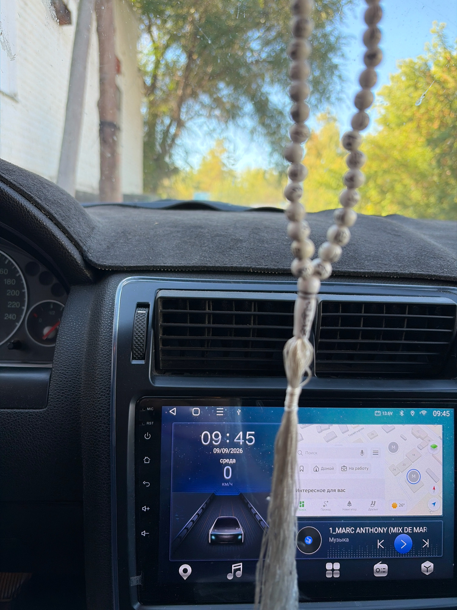 A medium shot captures the interior of a vehicle, focusing on the dashboard and a car infotainment system. A string of light-colored prayer beads hangs from the rearview mirror, obscuring part of the windshield and the view of trees and a building outside. The dashboard features a black textured surface and a black fabric cover. To the left, a speedometer dial is visible, showing a maximum reading of 240. The central infotainment screen displays a navigation map, current time (09:45), date (09/09/2026), and speed (0 km/h). Below the map, there are icons for search, home, work, and interesting suggestions. A music player interface is also visible, showing the song "1_MARC ANTHONY (MIX DE MAR)" and playback controls. The screen also shows battery voltage (13.6V) and signal strength indicators. The interior of the car is dark, contrasting with the brighter exterior view. The presence of the date "09/09/2026" suggests the image might be from the past or a simulation. The infotainment system suggests the vehicle is a modern sedan or SUV. The setting appears to be a residential area given the building in the background. The image is likely taken in the village of Zhelezinka, in the Zhelezinka rural district, Kazakhstan.