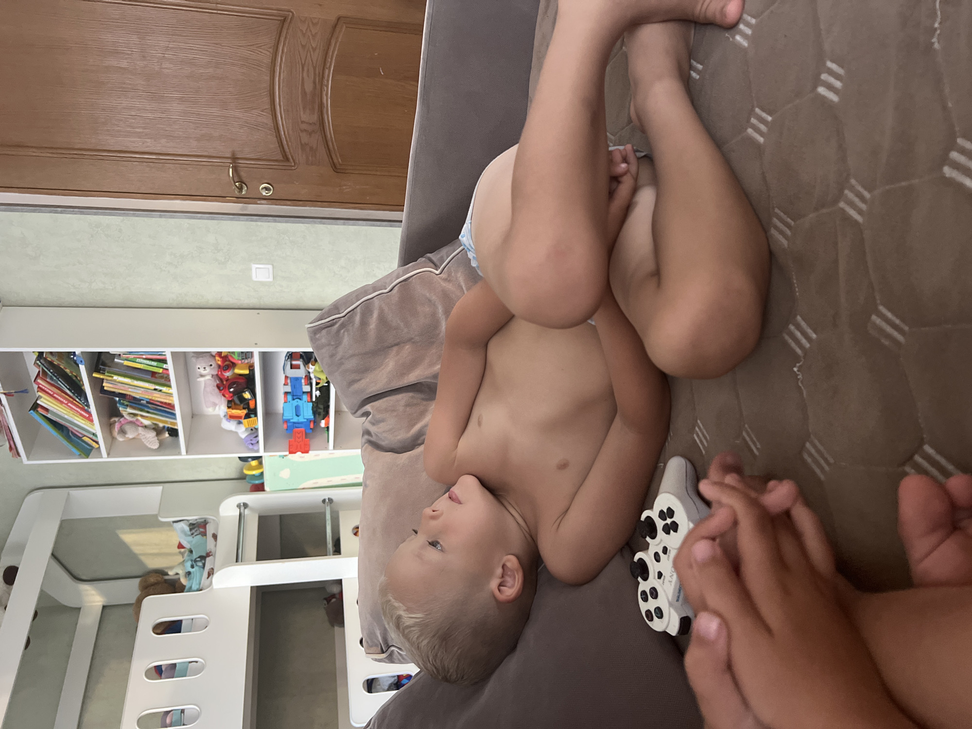 The image captures an indoor domestic scene, reportedly located in Sochi, Russia. A fair-skinned young male child, estimated to be between three and five years old, with short blonde hair and light eyes, is lying on his back on a brown upholstered sofa. He is shirtless, wearing only a light-colored diaper or training pants, and rests his head on a dark gray pillow. His gaze is directed upwards and to the right, beyond the immediate frame. A white PlayStation DualShock controller is positioned on the sofa cushion in the lower-center, near his left hand. In the foreground, the hands and feet of another individual, possibly a child, are partially visible, suggesting proximity to the boy.
To the left of the scene, a white, modern bunk bed with a curved design and a ladder is present. Bedding and stuffed animals are visible on the top bunk. To the right, a tall, white bookshelf is filled with a variety of children's books, toys, and stuffed animals. The wall behind the bookshelf is a light, textured greenish-beige. A white electrical outlet or light switch is mounted on this wall. Further to the right, a light brown wooden door with a brass handle and a rounded panel design is partially visible, indicating an exit to another room or hallway.