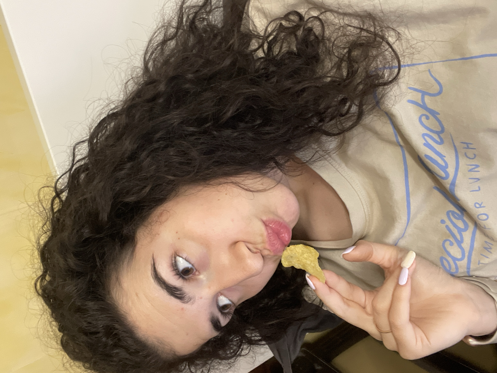 A young woman with curly dark hair is holding a potato chip towards her open mouth. She is wearing a beige top with light blue writing. Her fingernails are painted in a pale pink and white style. The background is a plain wall and countertop. The location in Sofia, Bulgaria is not discernible from the image.